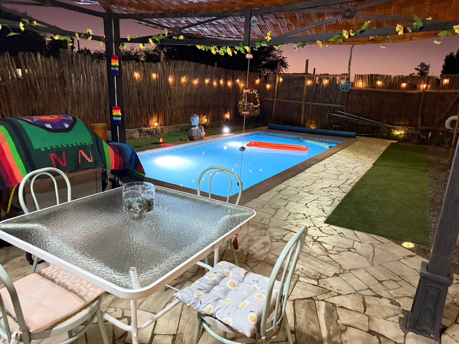 Patio con piscina iluminada, mesa con fuente, tumbonas y pérgola al atardecer.