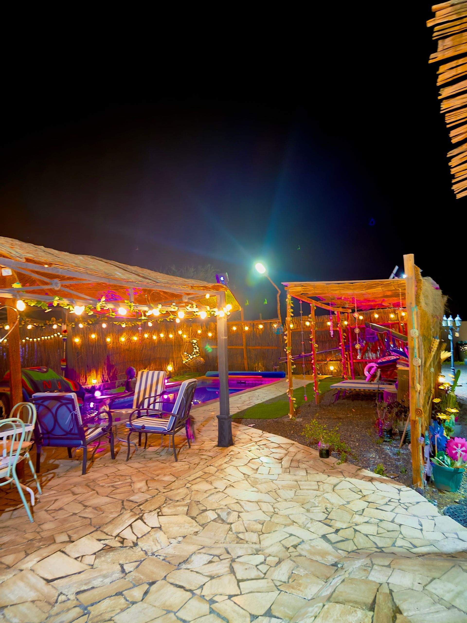 Zona de comedor al aire libre colorida y cálidamente iluminada con guirnaldas de luces y mesas bajo pérgolas por la noche.