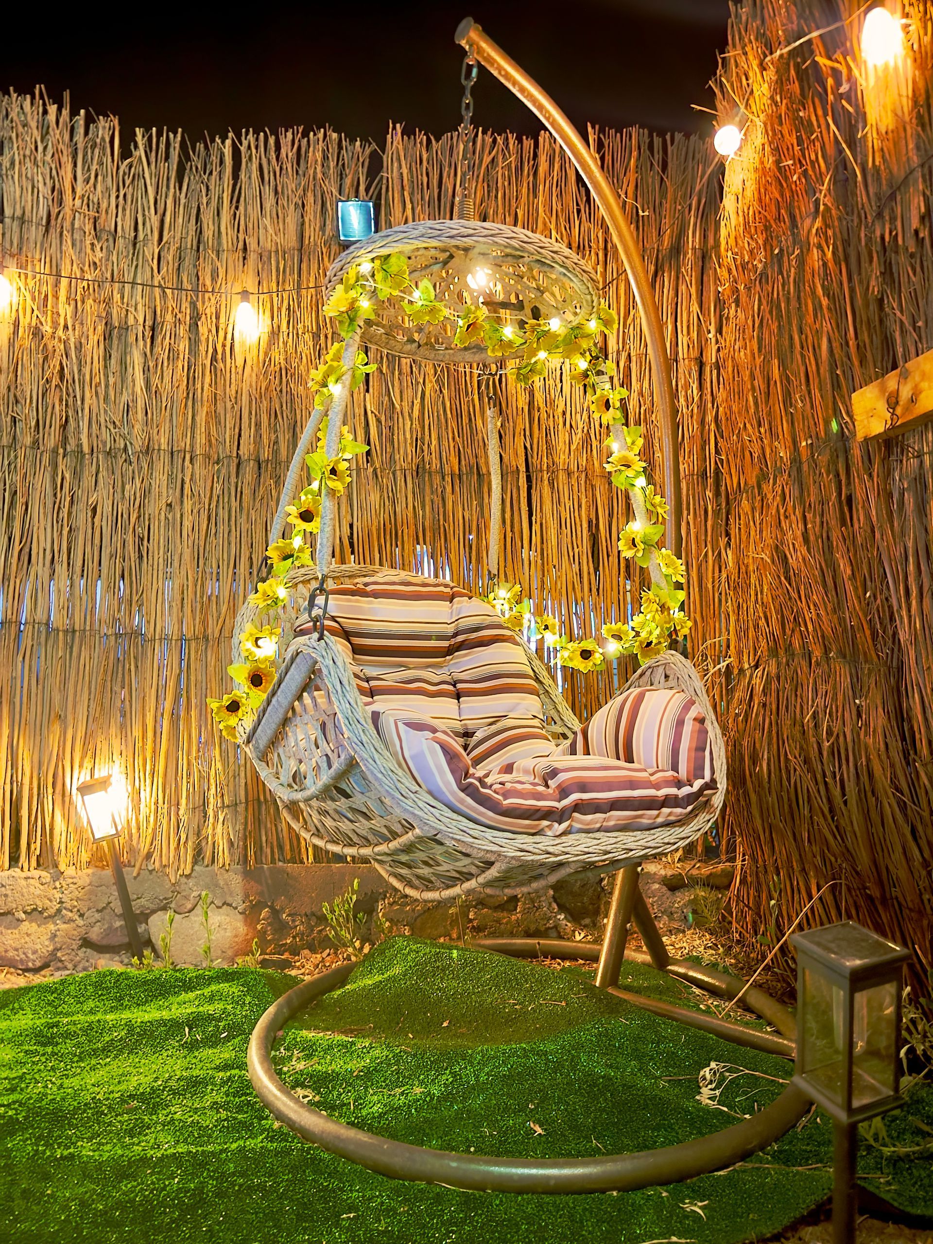Silla colgante decorativa con forma de huevo sobre un césped verde con luces de guirnalda y un fondo de bambú por la noche.