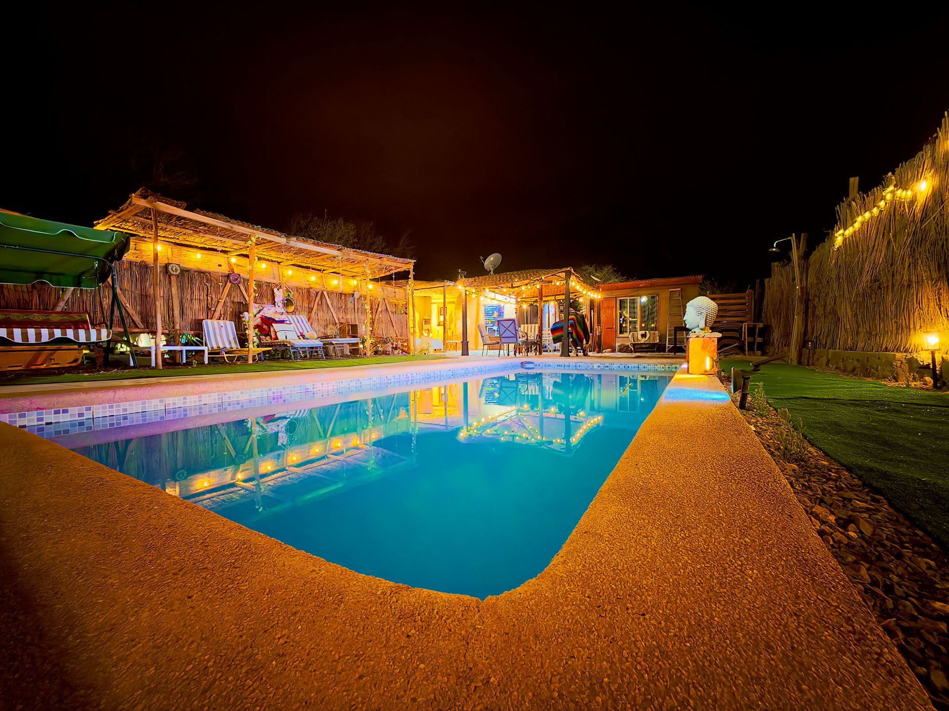 Piscina nocturna del complejo iluminada en azul y dorado, con cabañas y palmeras a lo largo de la terraza.