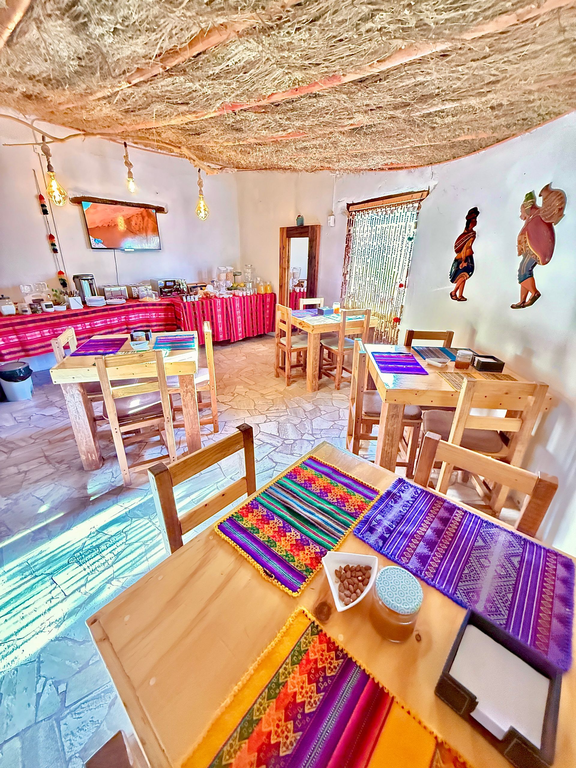Comedor rústico y luminoso con mesas de madera, coloridos caminos de mesa tejidos y un largo banco rosa junto a la pared.