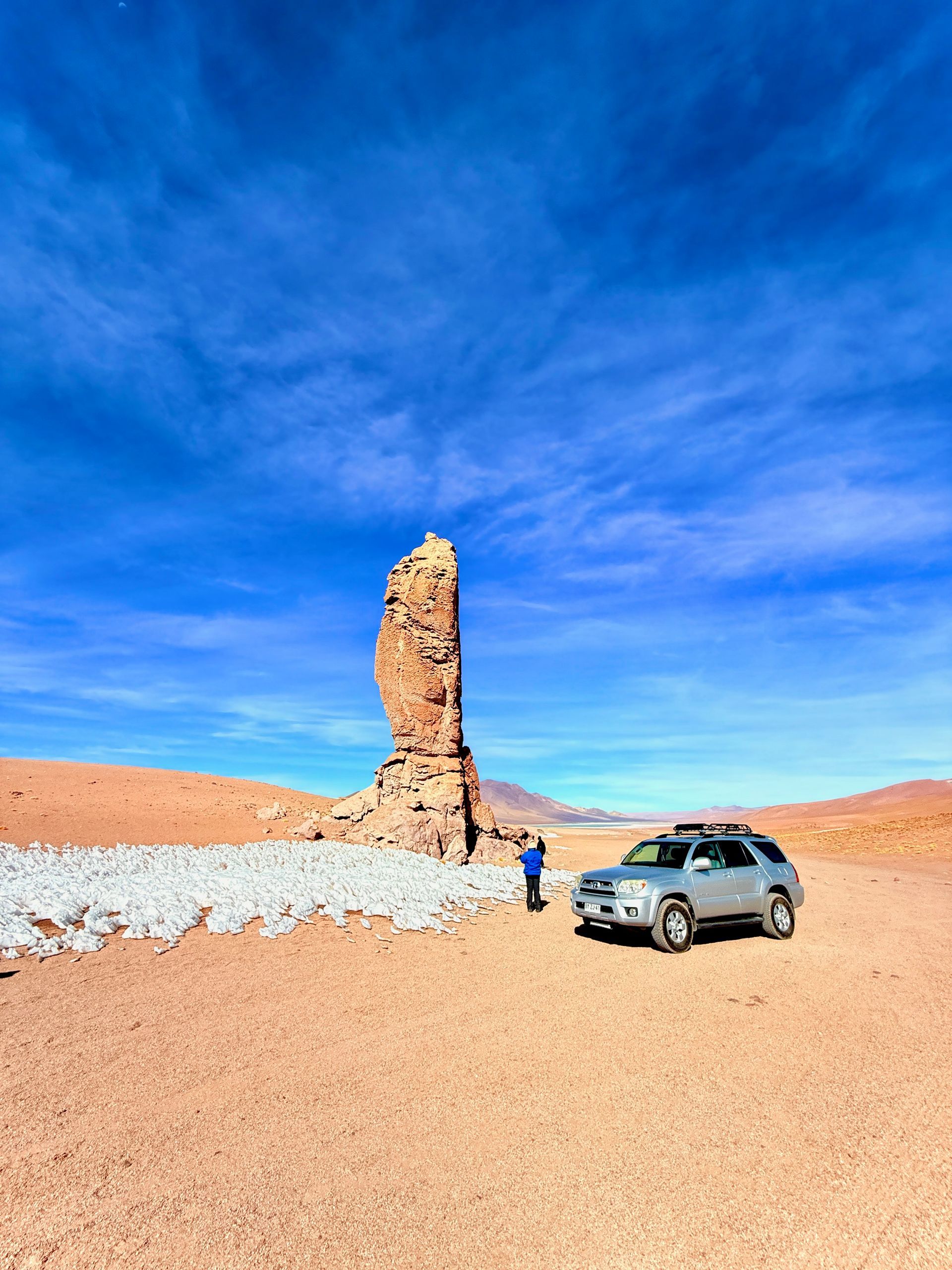 Un todoterreno y dos personas junto a una alta aguja rocosa en un desierto bajo un cielo azul brillante.