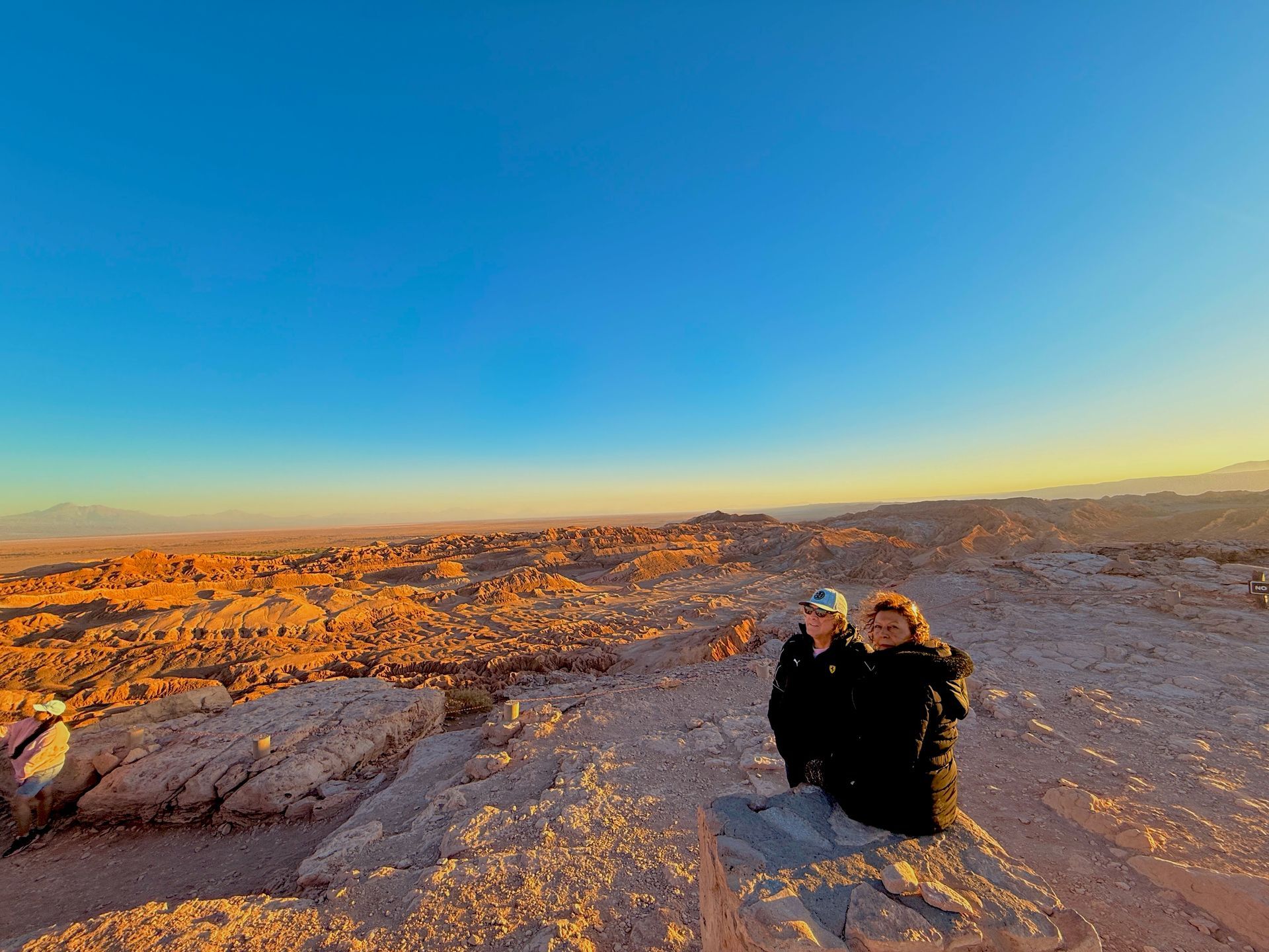 Dos excursionistas sentados en una cresta rocosa del desierto al atardecer, bajo un amplio cielo azul anaranjado.