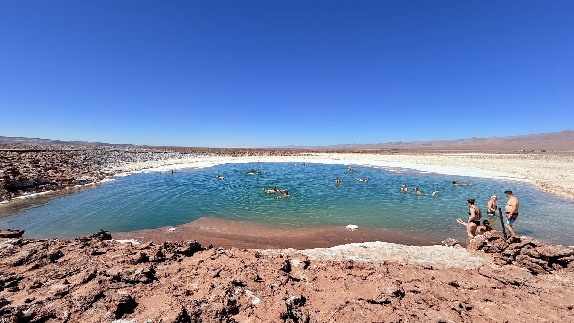 Laguna turquesa en un paisaje desértico rocoso con algunas personas de pie en la orilla.
