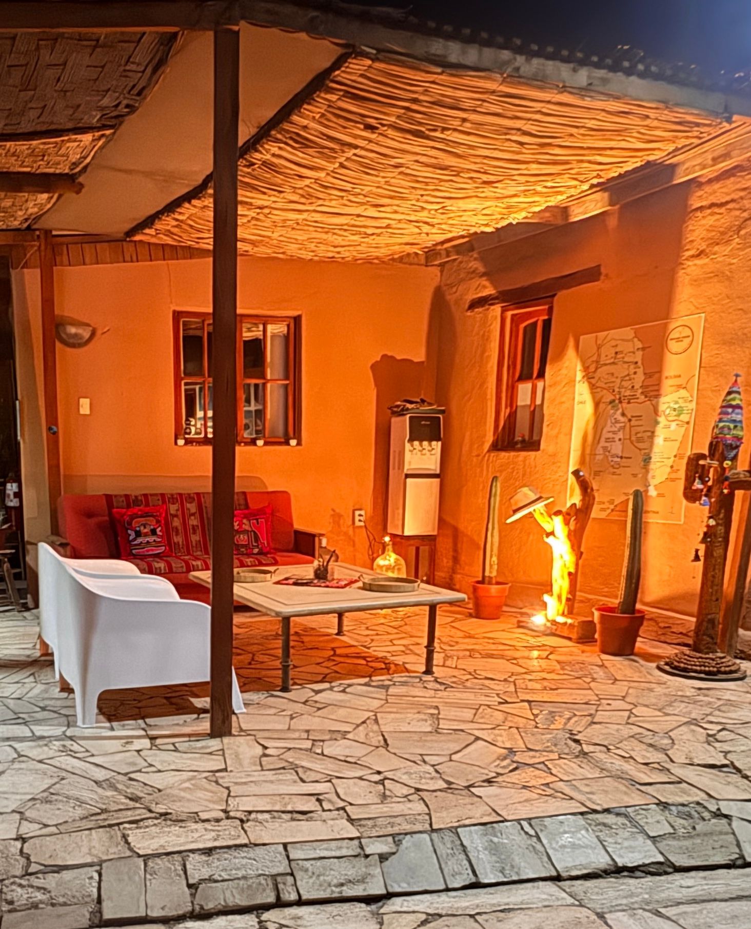 Patio con iluminación cálida, suelo de piedra, asientos rojos y un calefactor encendido bajo un techo de paja.