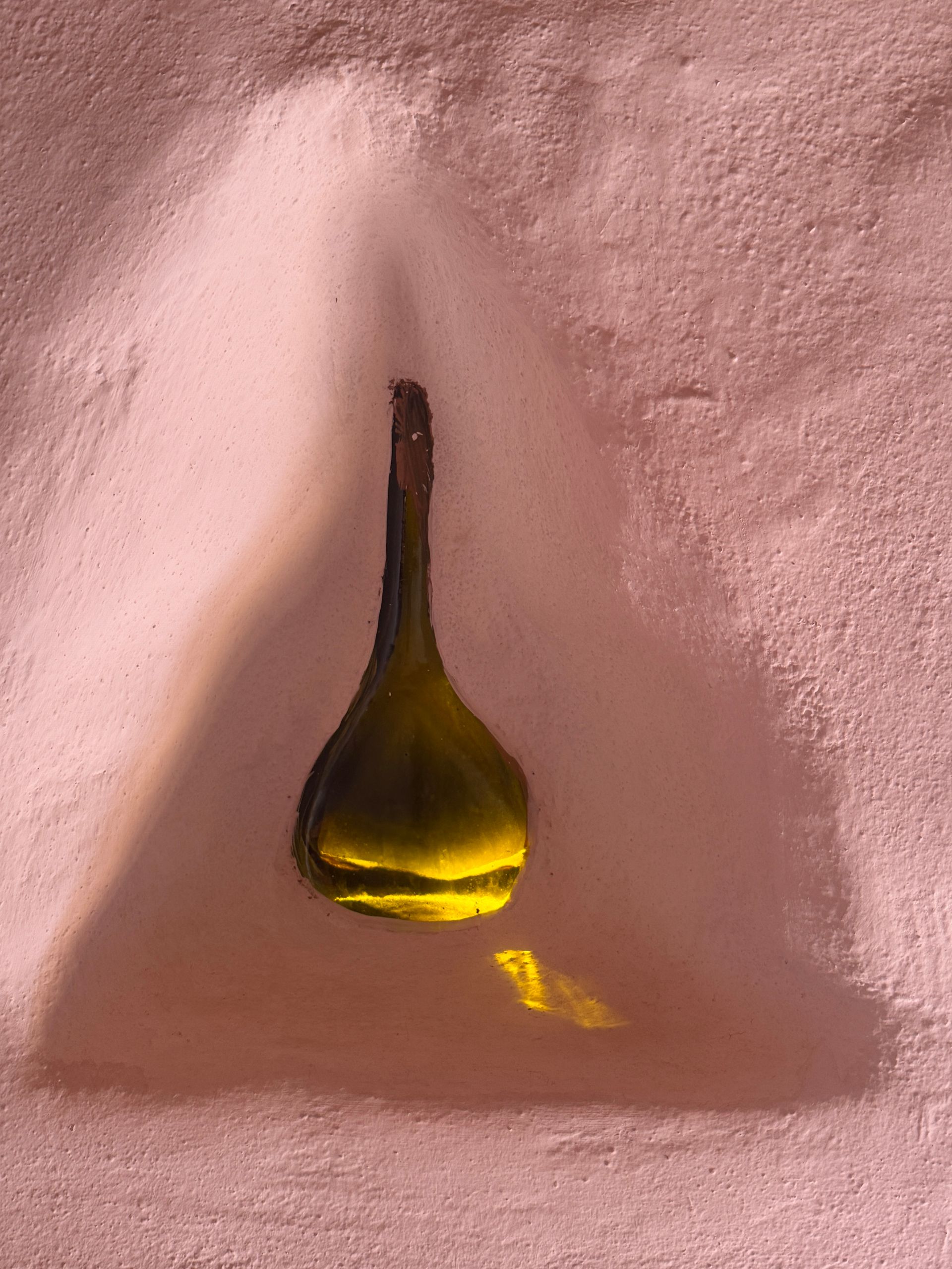 Lámpara colgante de cristal amarillo sobre una pared rosa texturizada, iluminada desde arriba.