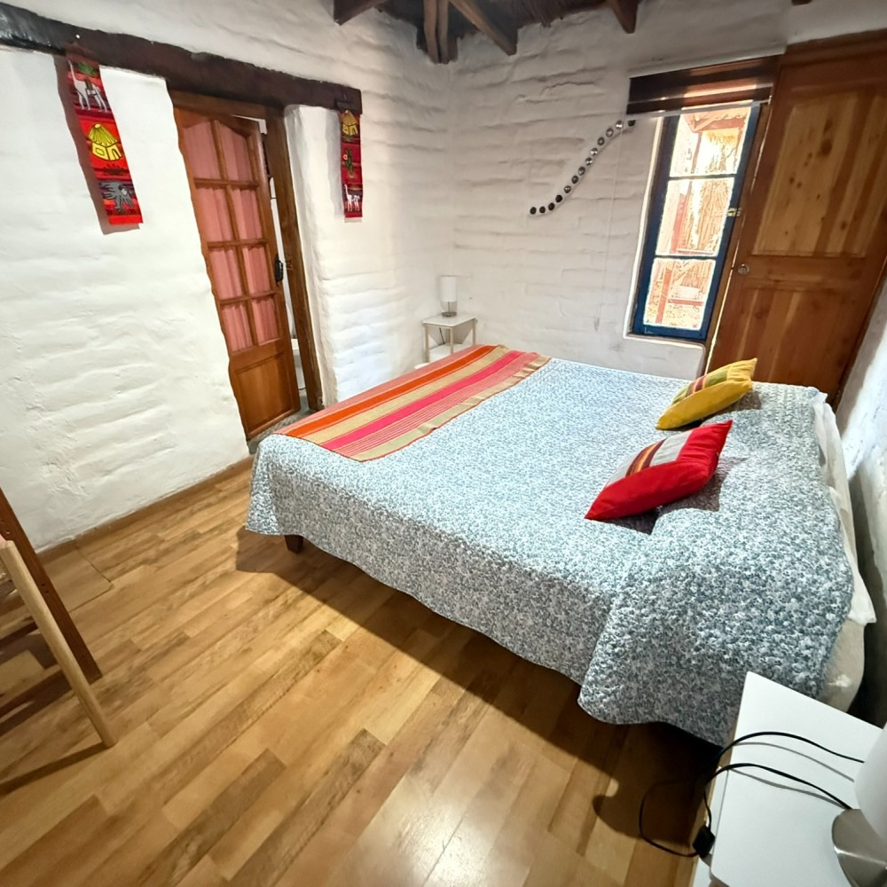 Acogedora habitación con cama, suelo de madera, paredes de ladrillo blanco y cojines coloridos cerca de dos ventanas.