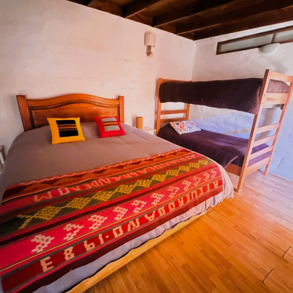 Habitación con cama de matrimonio, litera y coloridas mantas tejidas sobre suelo de madera.