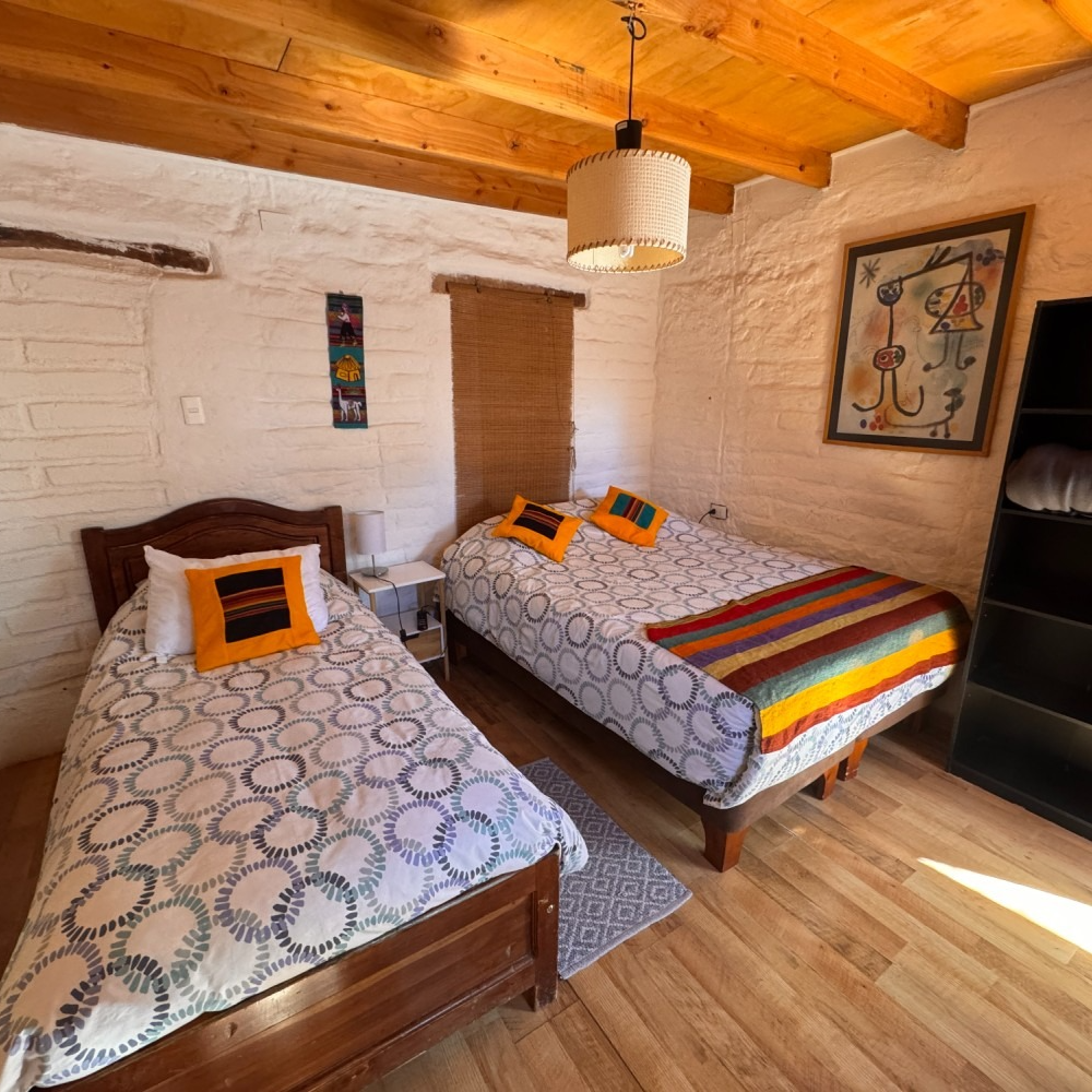 Habitación acogedora con dos camas individuales, ropa de cama estampada, techo de madera y luz natural cálida.