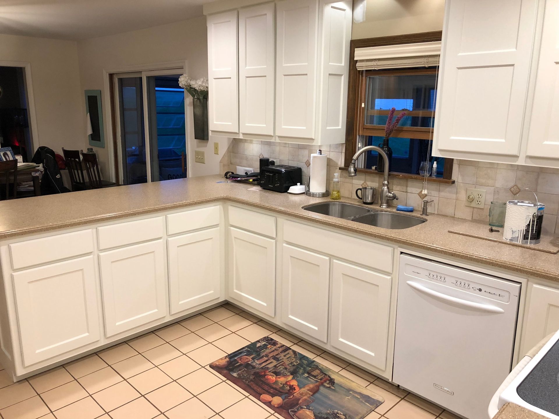 White kitchen cabinets and appliances, beige countertops, tile floor, sink by window.