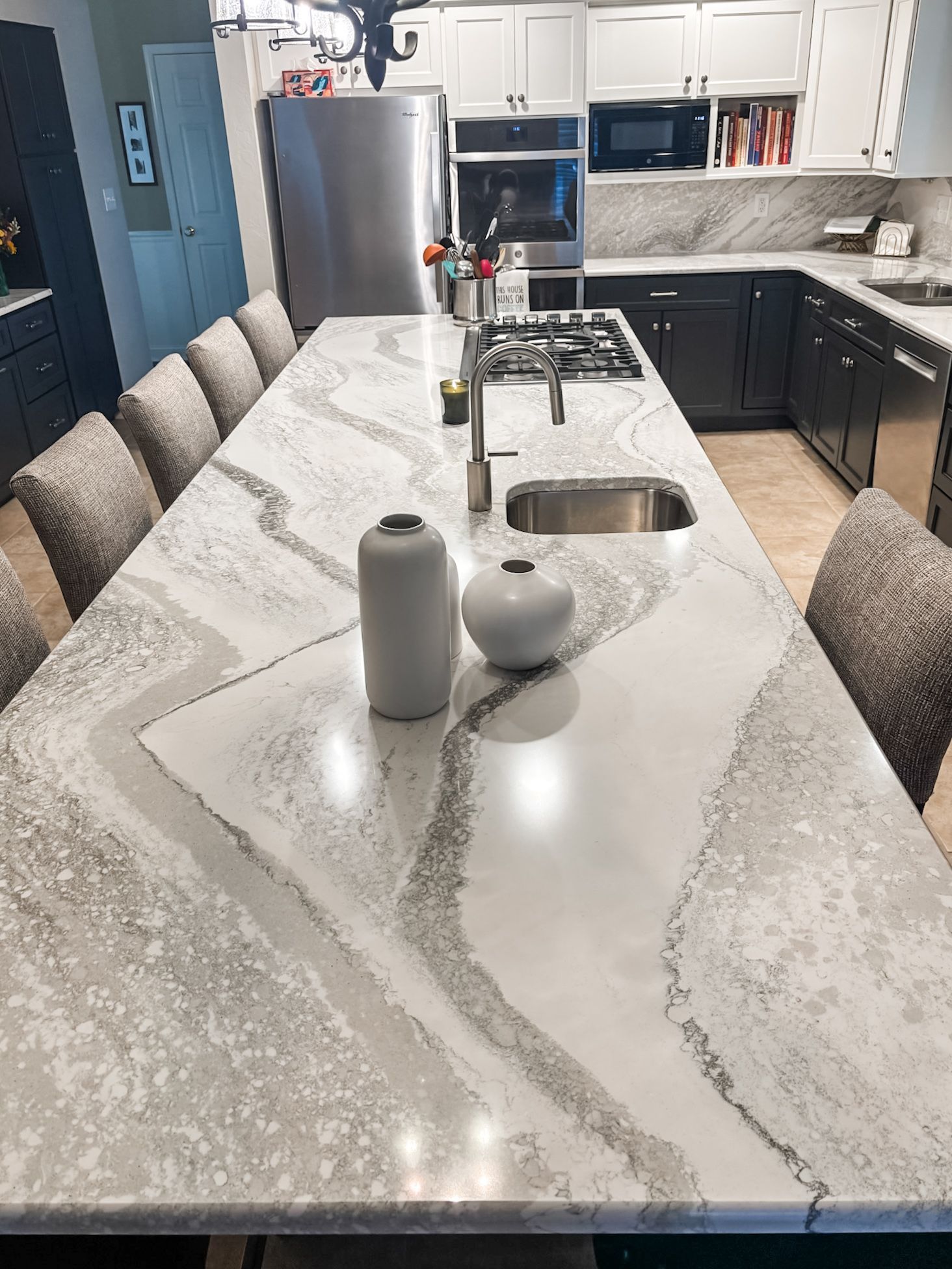 Modern kitchen with a long white countertop island with seating.  Stainless steel appliances and gray cabinets.