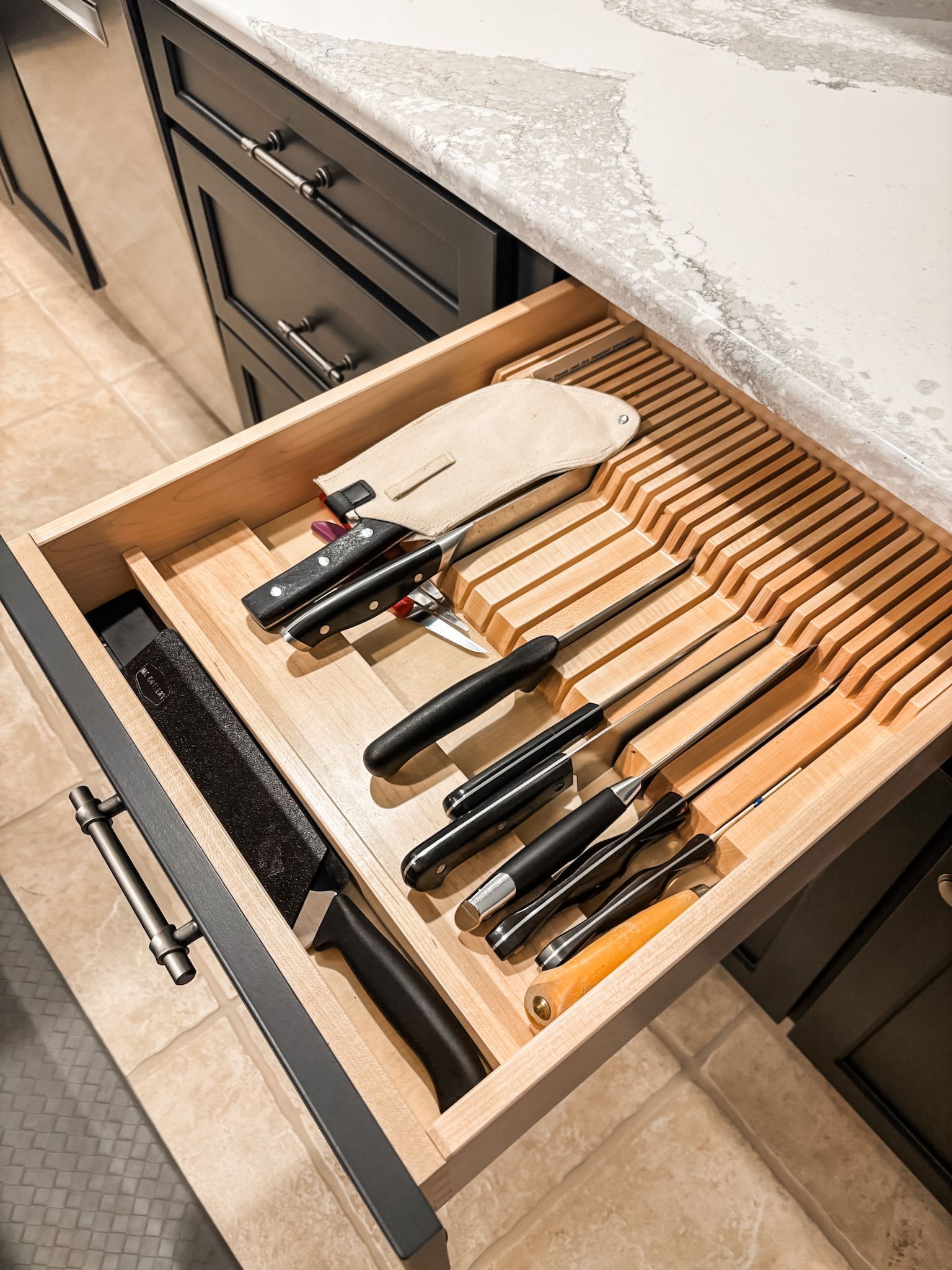 Open kitchen drawer with knives in a wooden organizer. Black handles, light countertop, dark cabinets.