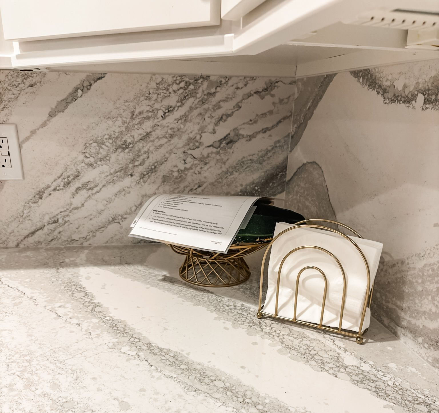 Kitchen counter with white and gray marbled countertop. A cookbook on a gold pedestal and a napkin holder are visible.