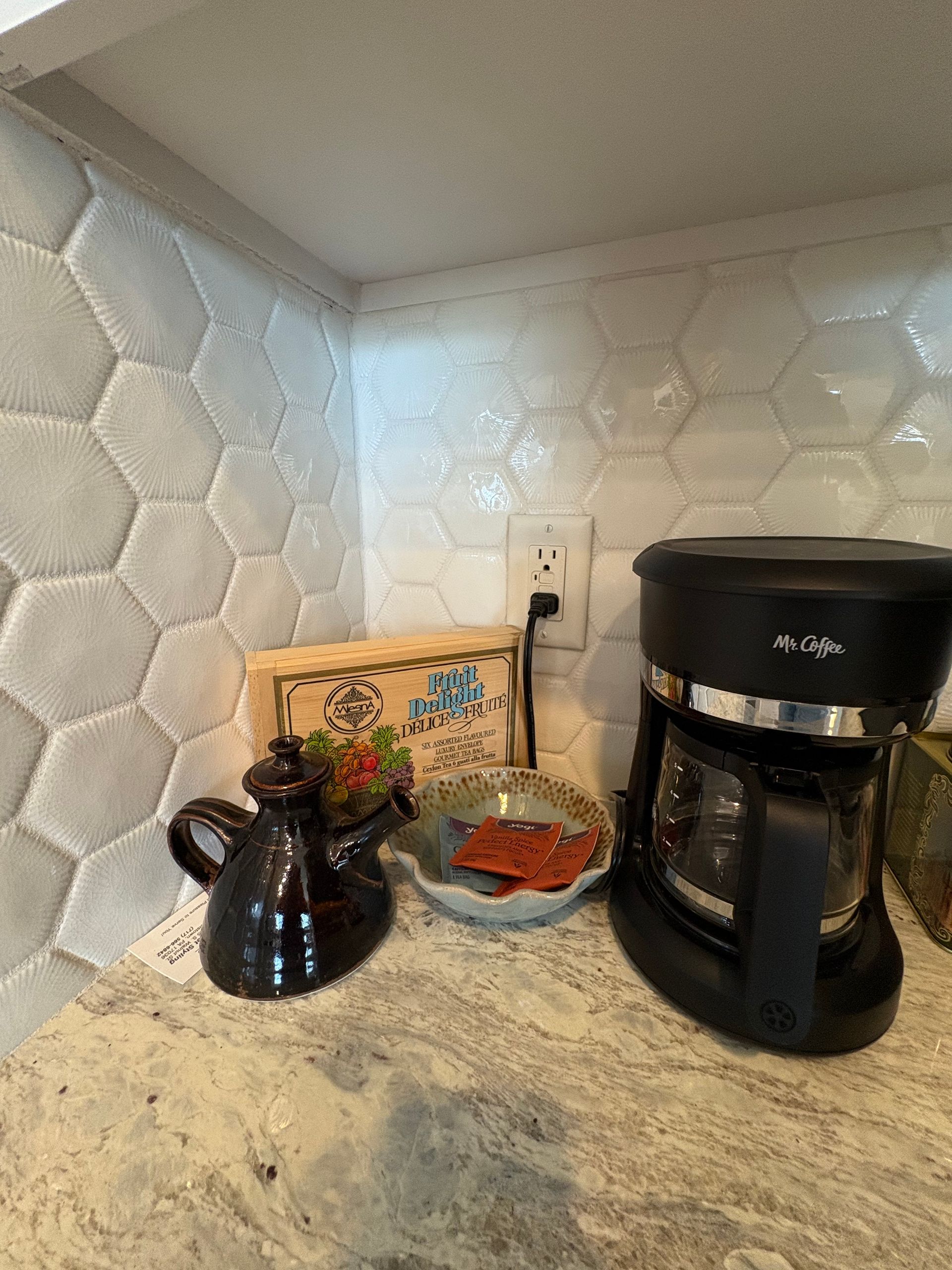 Coffee maker, ceramic pitcher, and decorations on a countertop with hexagonal tiled backsplash.