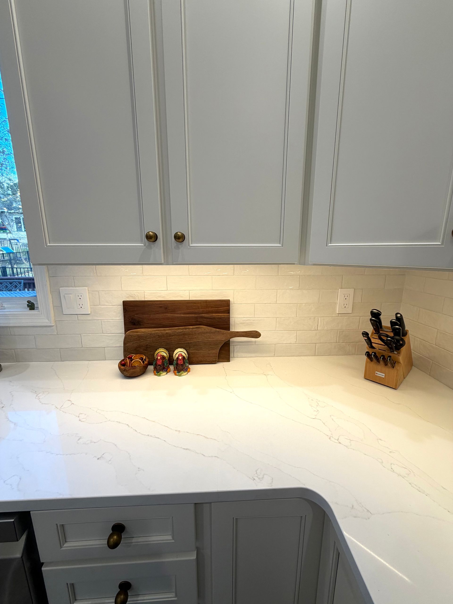 Kitchen countertop with wooden cutting board, small figurines, and knife block under white cabinets.