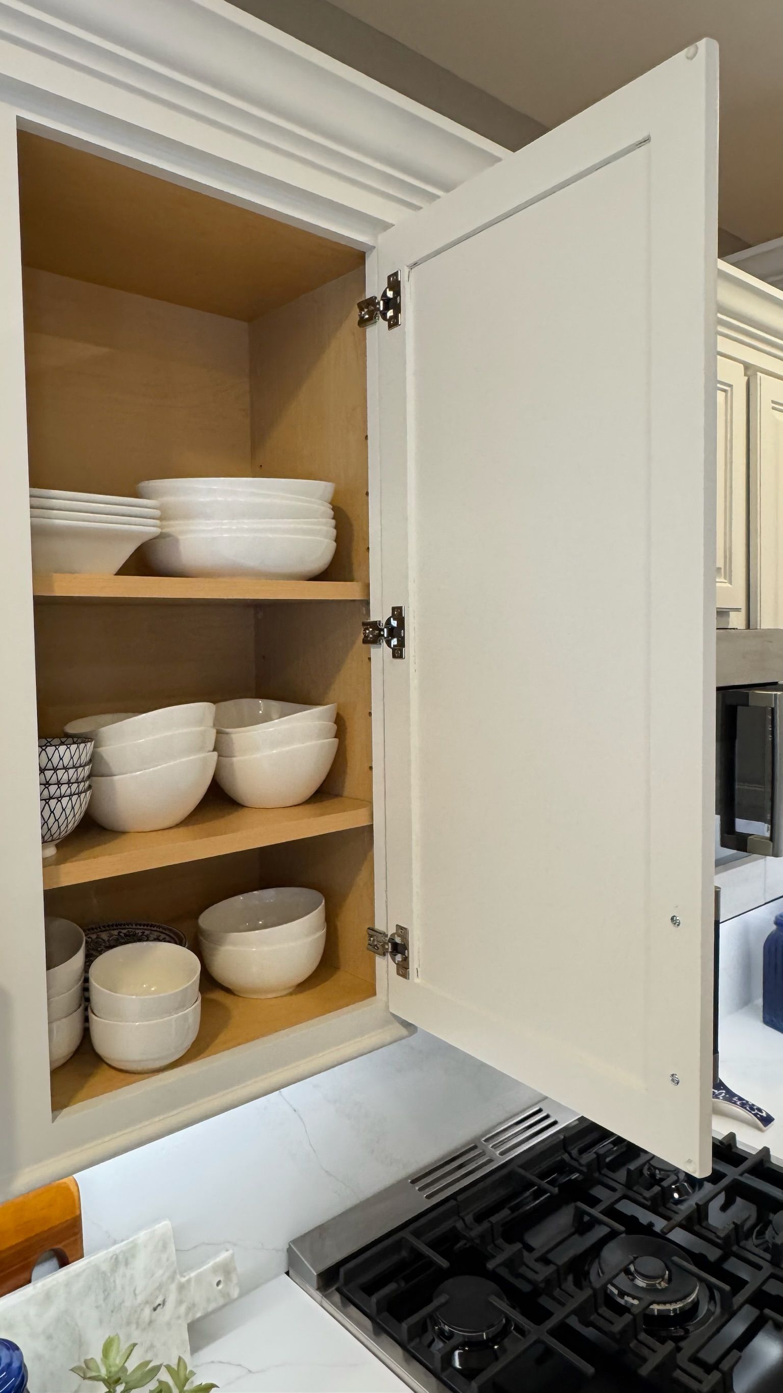 White kitchen cabinet open, revealing stacks of white bowls on wooden shelves, above a stovetop.