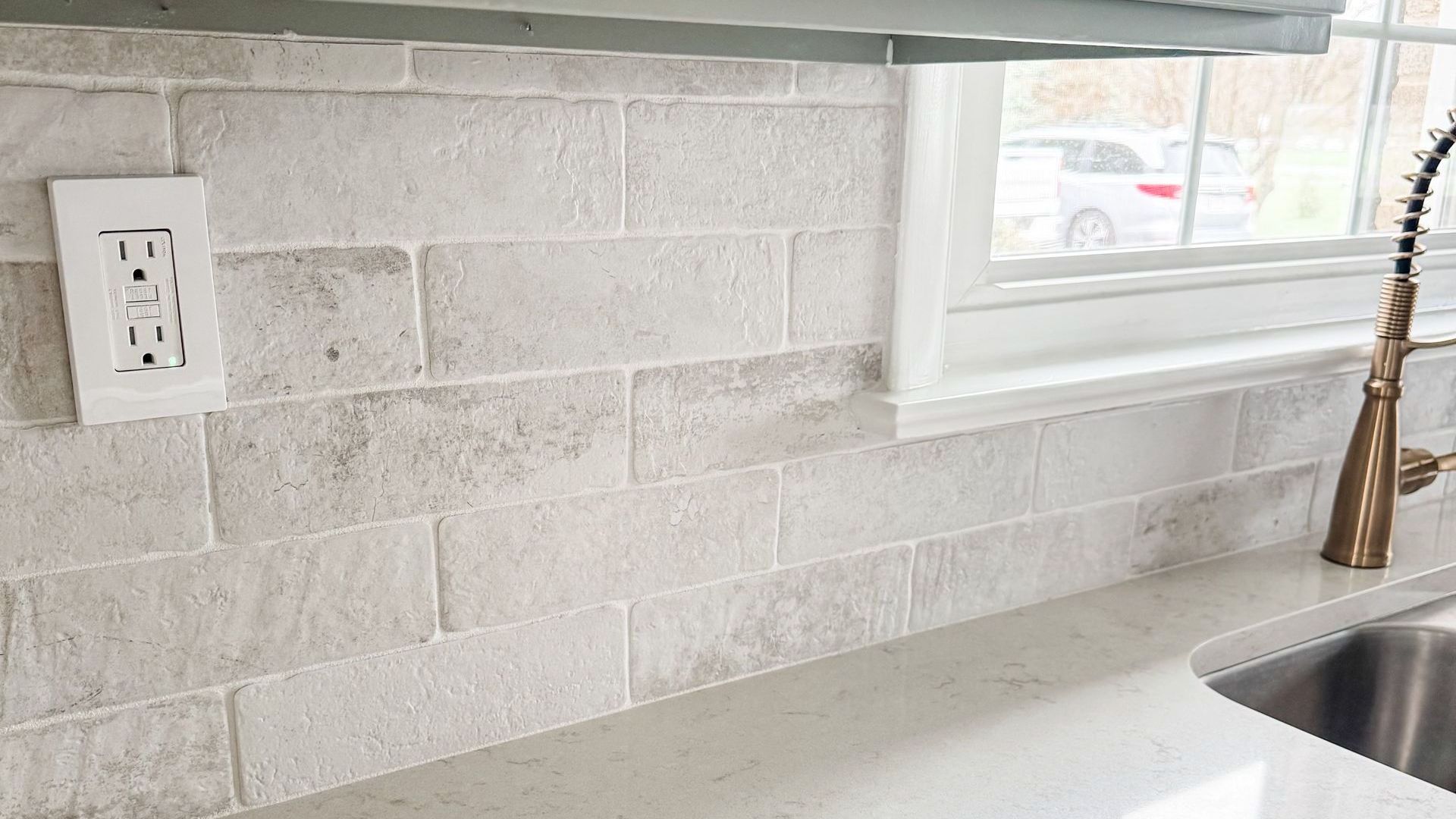 White brick-patterned backsplash above a light countertop with a window and faucet.