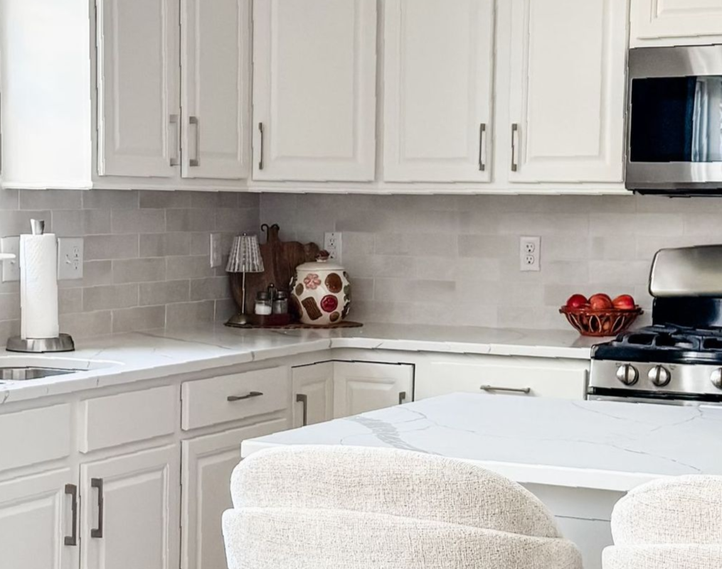 White kitchen with countertops, cabinets, and appliances. Includes a countertop with fruit and a kitchen island with stools.