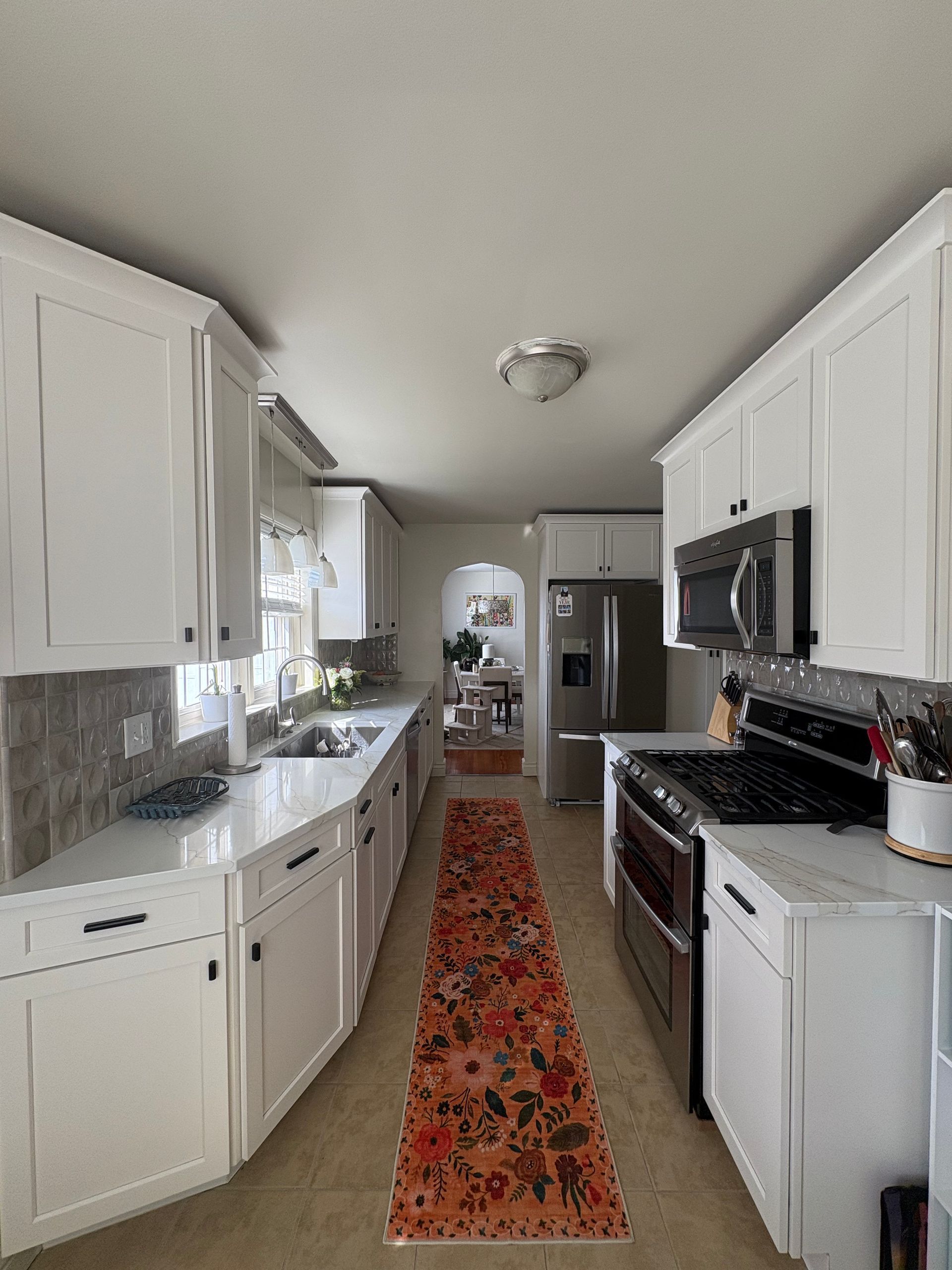 Long, white kitchen with a floral runner, stainless steel appliances, and a view into a dining area.