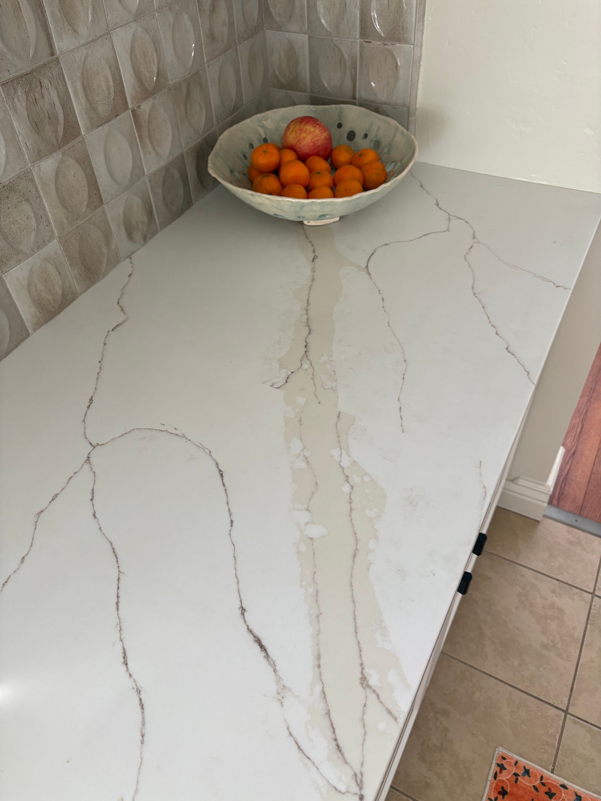 White countertop with marble-like veining, bowl of fruit, tiled backsplash.