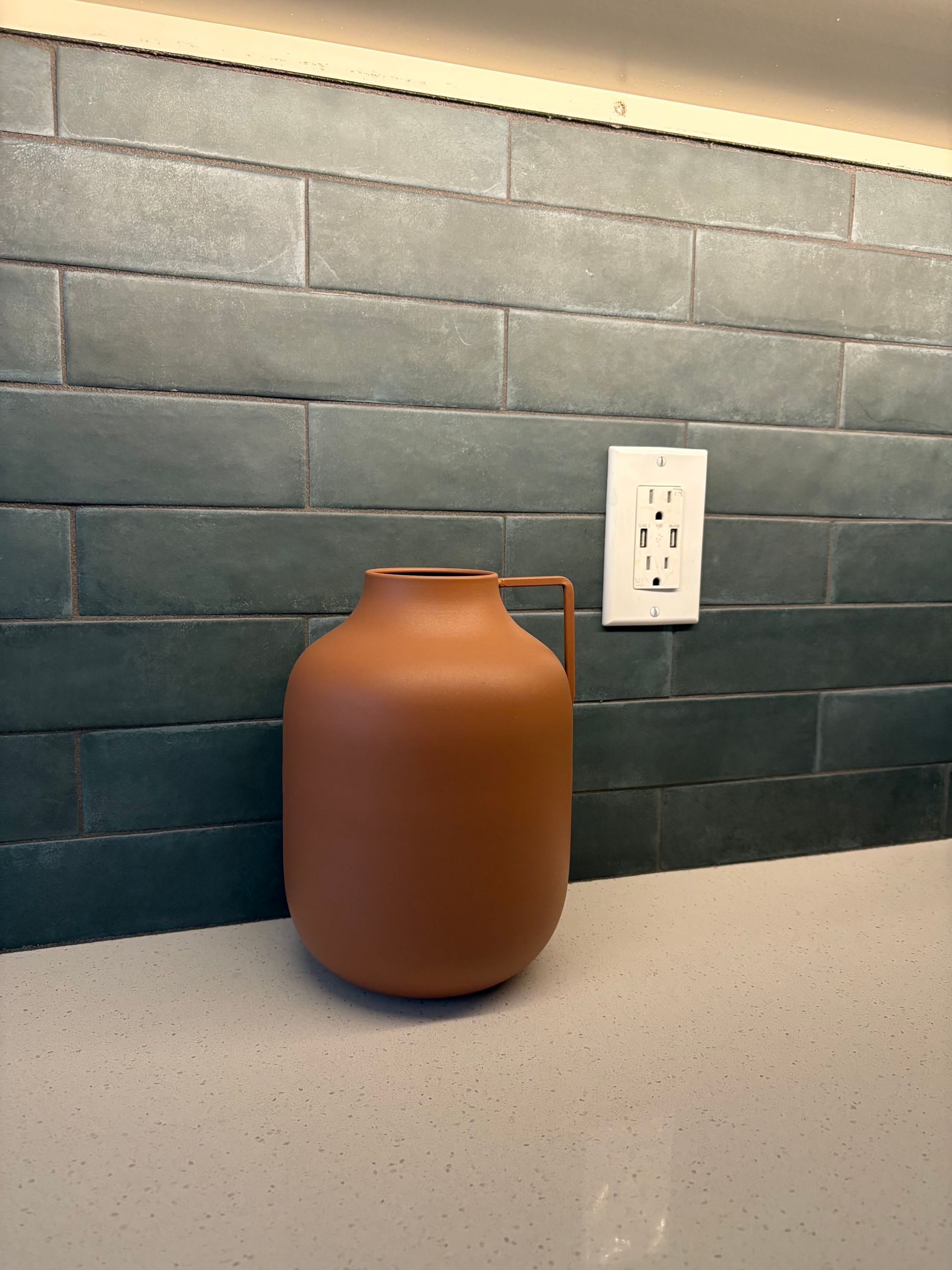 Brown ceramic vase on a light-colored countertop in front of a blue-tiled wall with an electrical outlet.