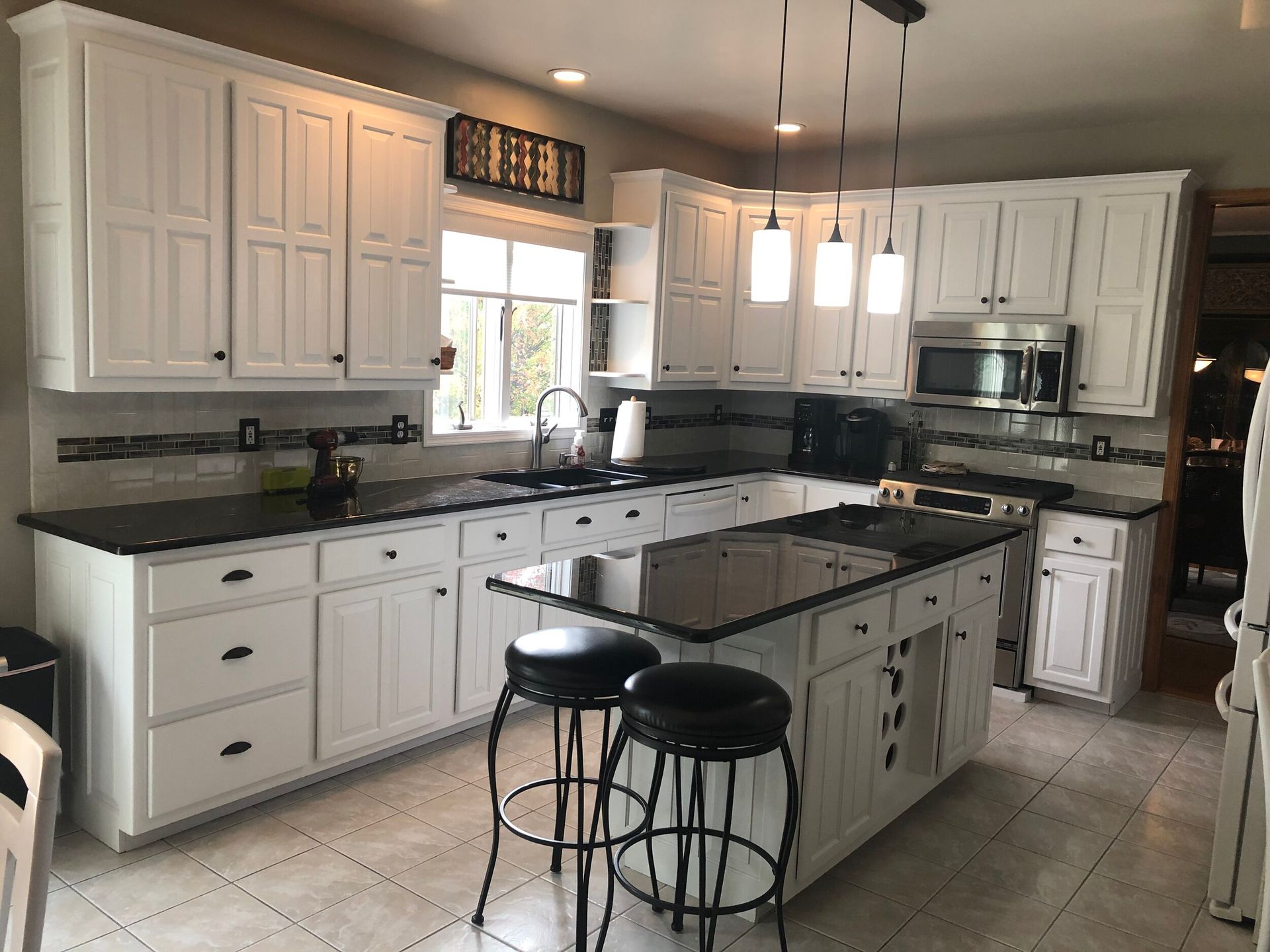 White kitchen with black countertops and island, featuring stools, overhead lights, and a window.
