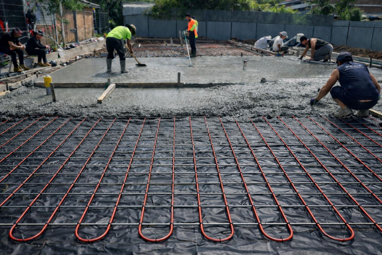 Construction workers pouring concrete over red underfloor heating pipes on a foundation slab
