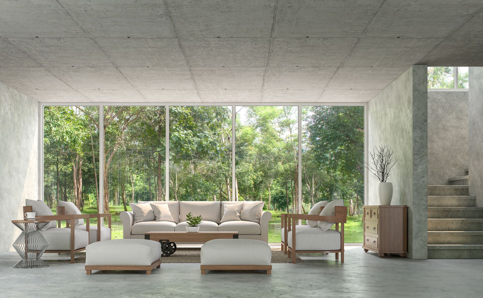 Modern living room with large windows overlooking trees; beige furniture, concrete walls, stairs.