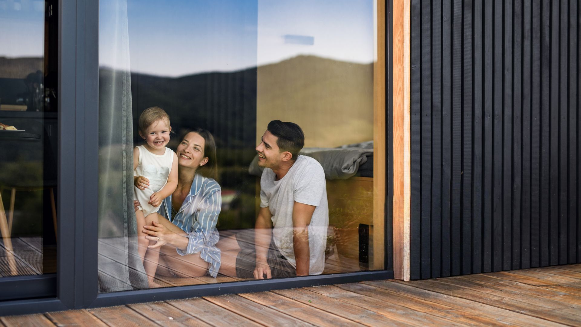 Family of three smiling while looking through a window. Wooden deck and dark paneling are visible.