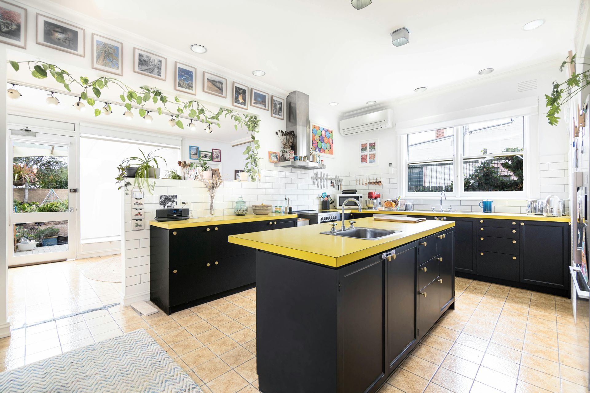 Bright kitchen with black cabinets, yellow countertops, white tile floor, and many windows and plants