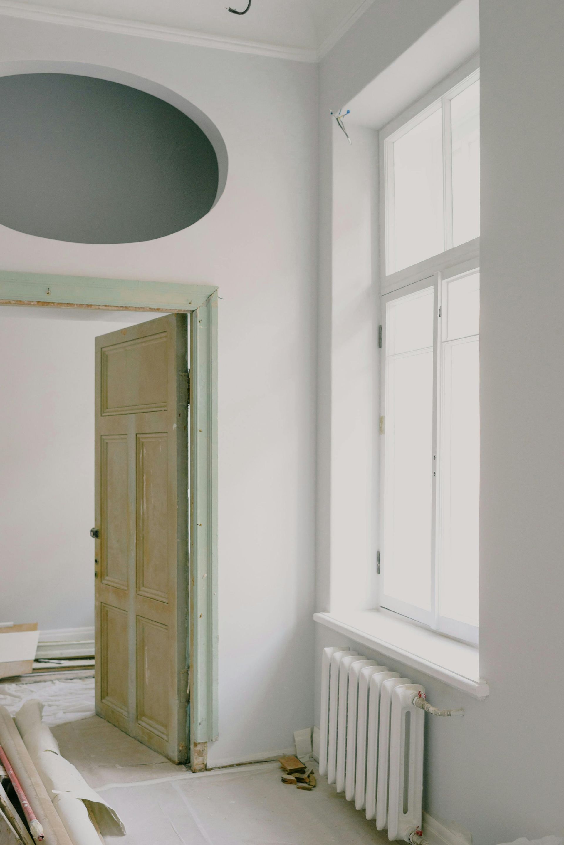 Bright white room with open wooden door, tall window, radiator, and oval ceiling opening.