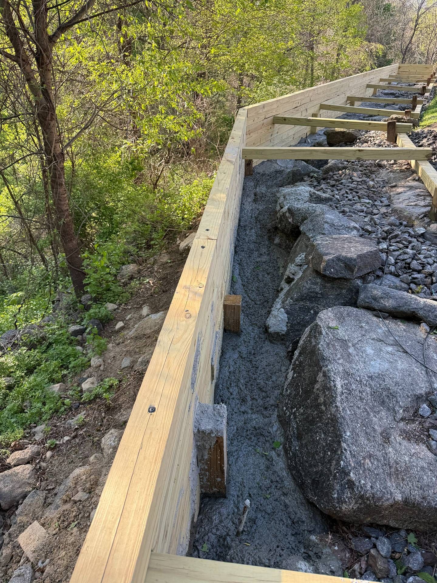 Wooden boardwalk under construction beside large rocks and green forest vegetation