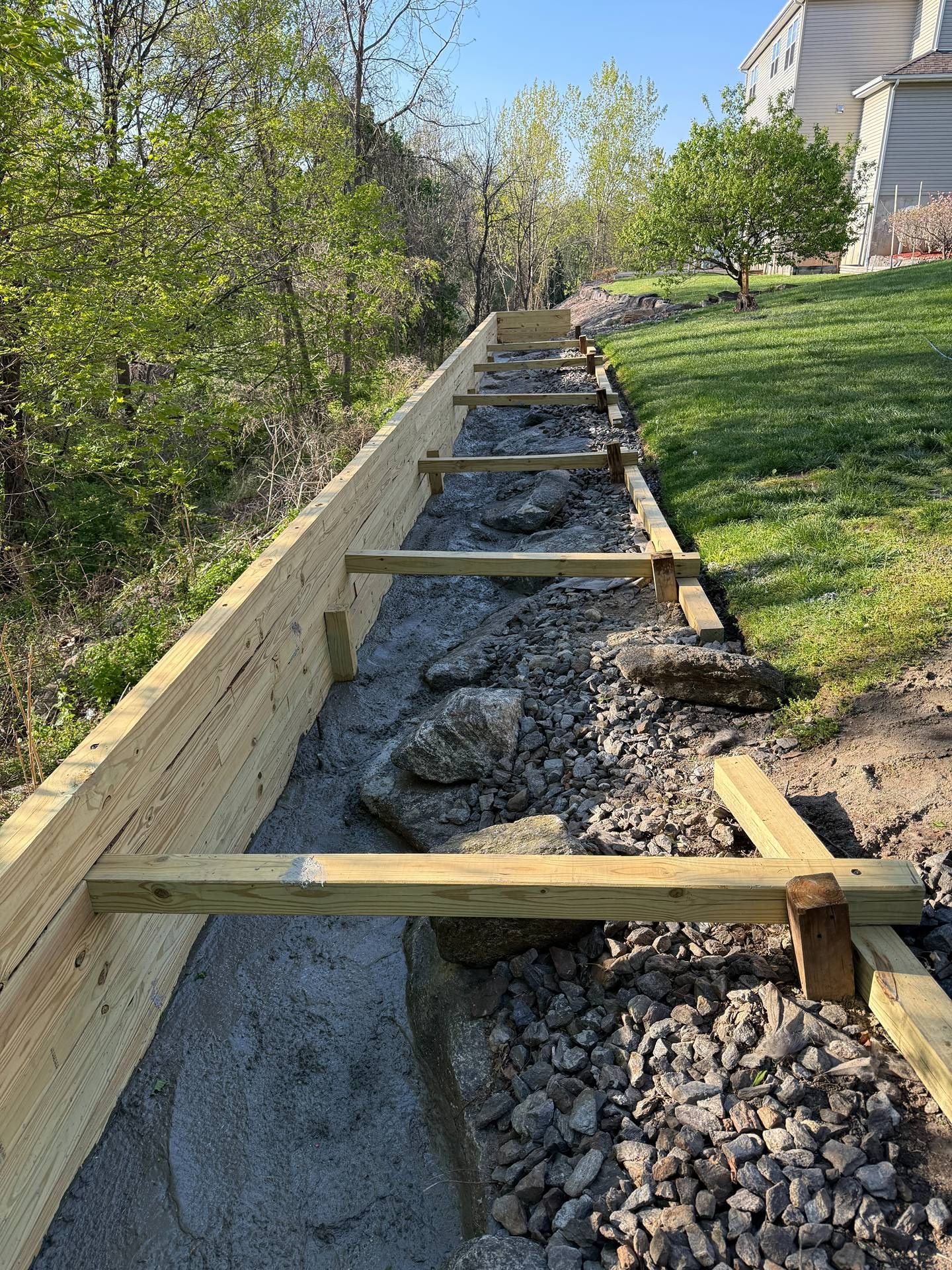 Wooden retaining wall supporting a gravel drainage trench beside a grassy hillside and house