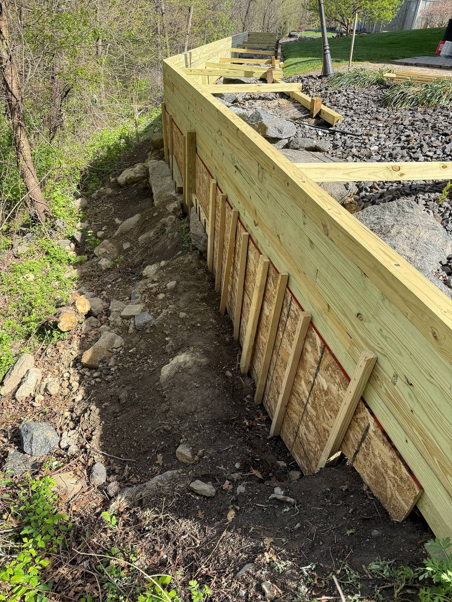 Wooden retaining wall under construction beside a dirt slope with gravel and beams on top.