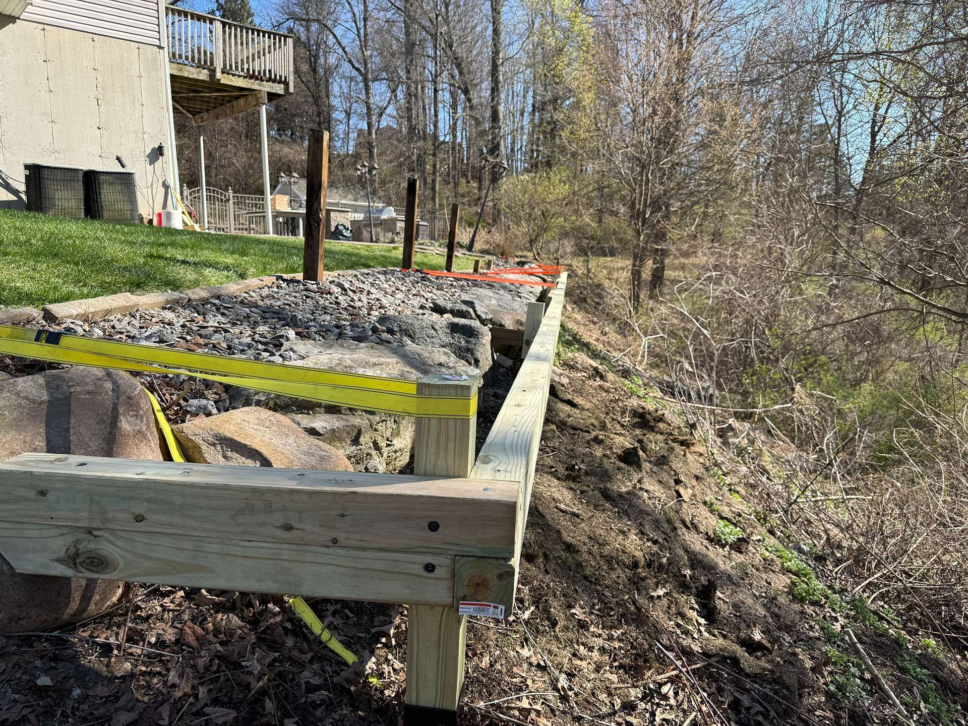 Wooden raised garden bed with black tarp and yellow tape beside a house, overlooking a wooded slope