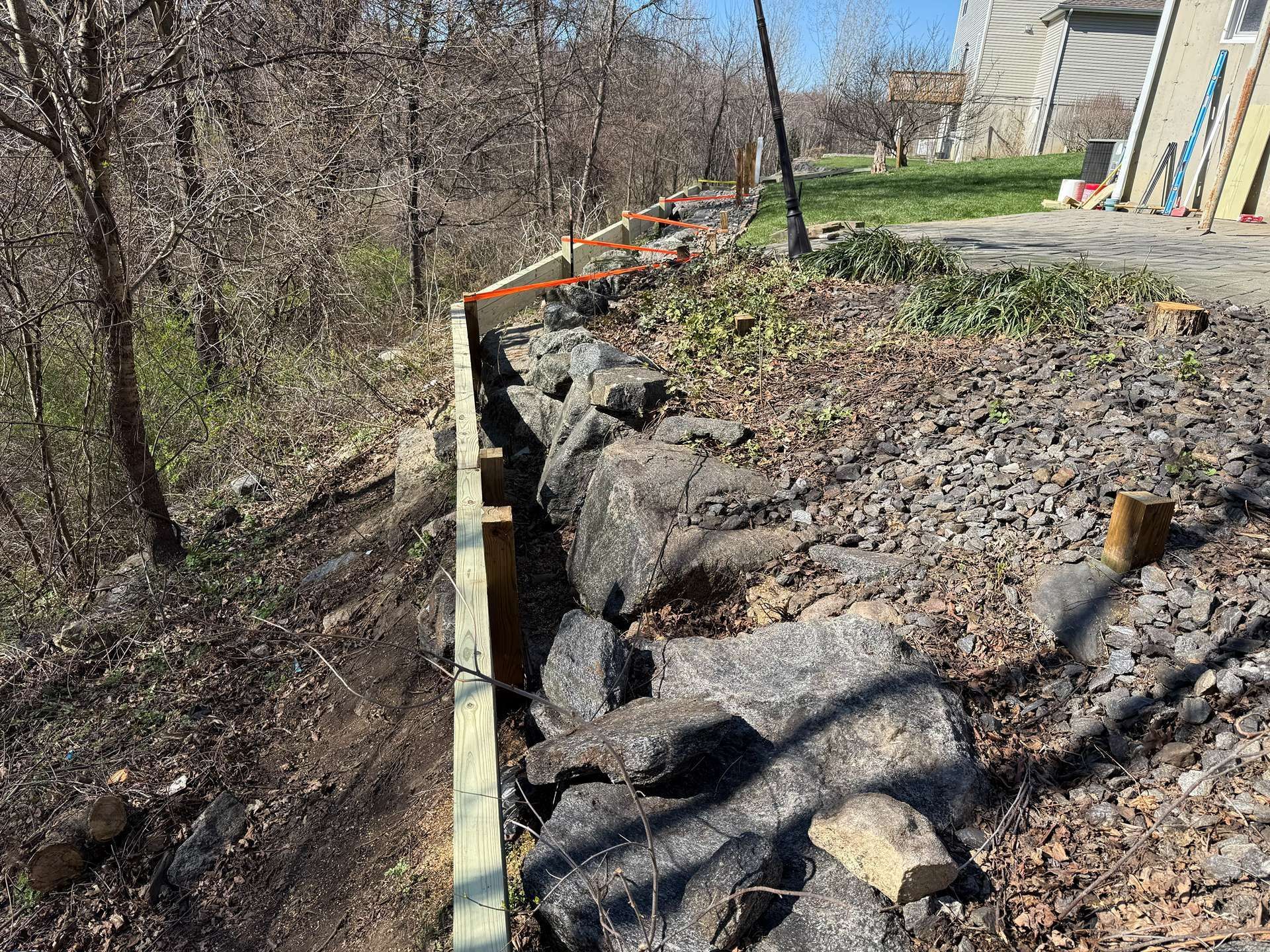 Retaining wall under construction beside a house, with rocks, gravel, and orange safety fencing