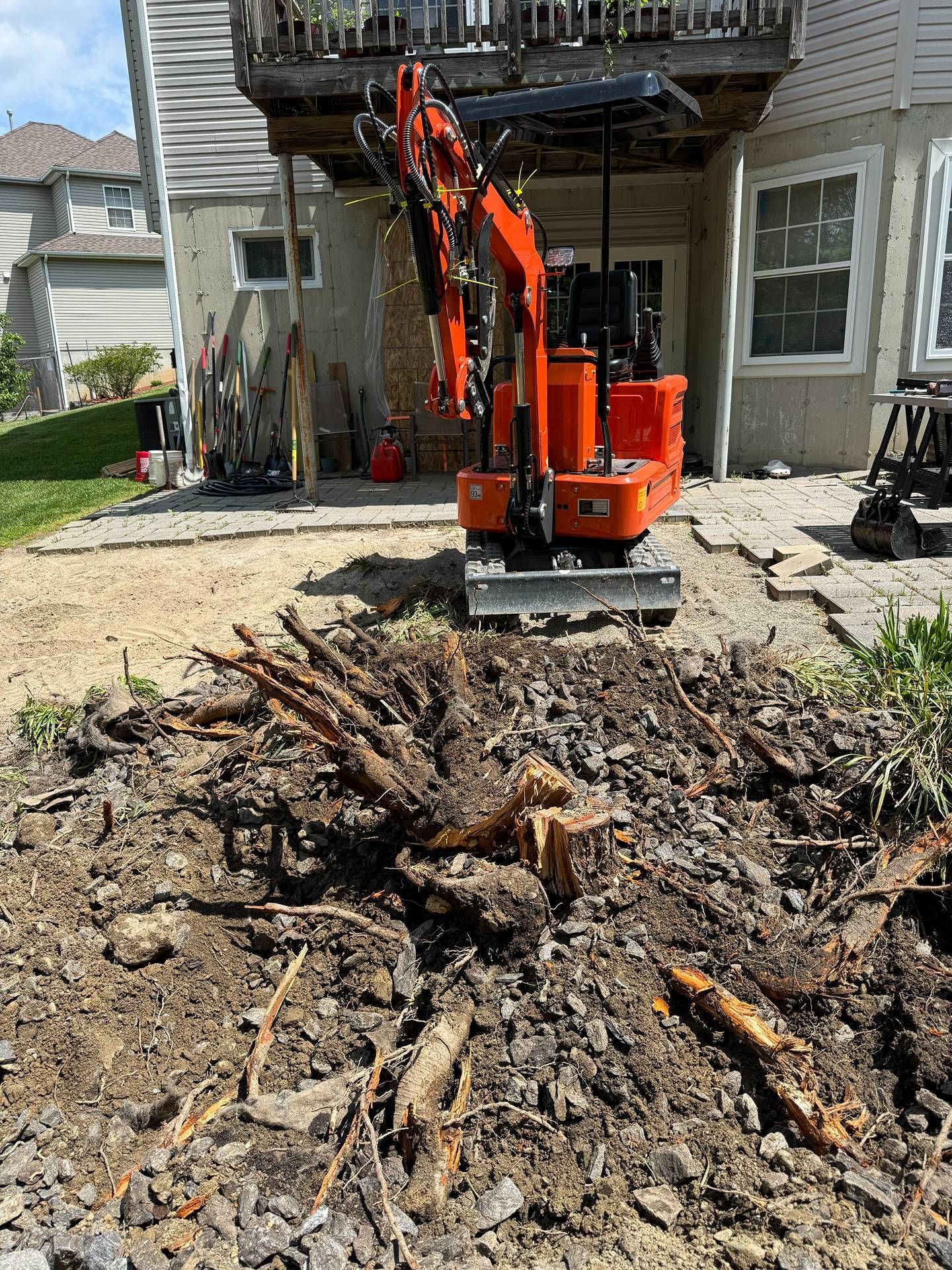 Orange excavator uprooting a large tree stump in a backyard beside a house