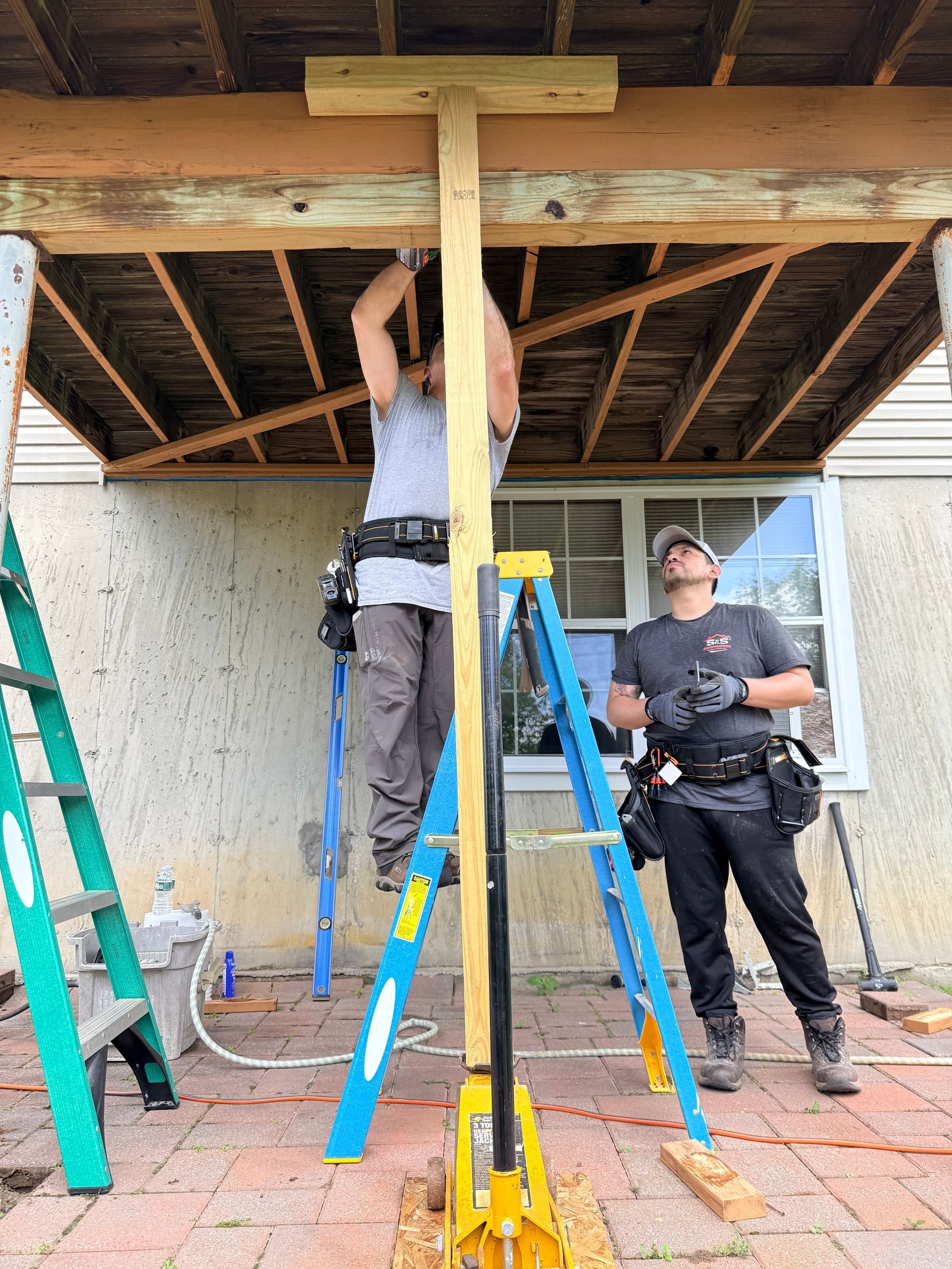 Two workers on ladders inspecting a porch ceiling with exposed beams and tools on the ground.