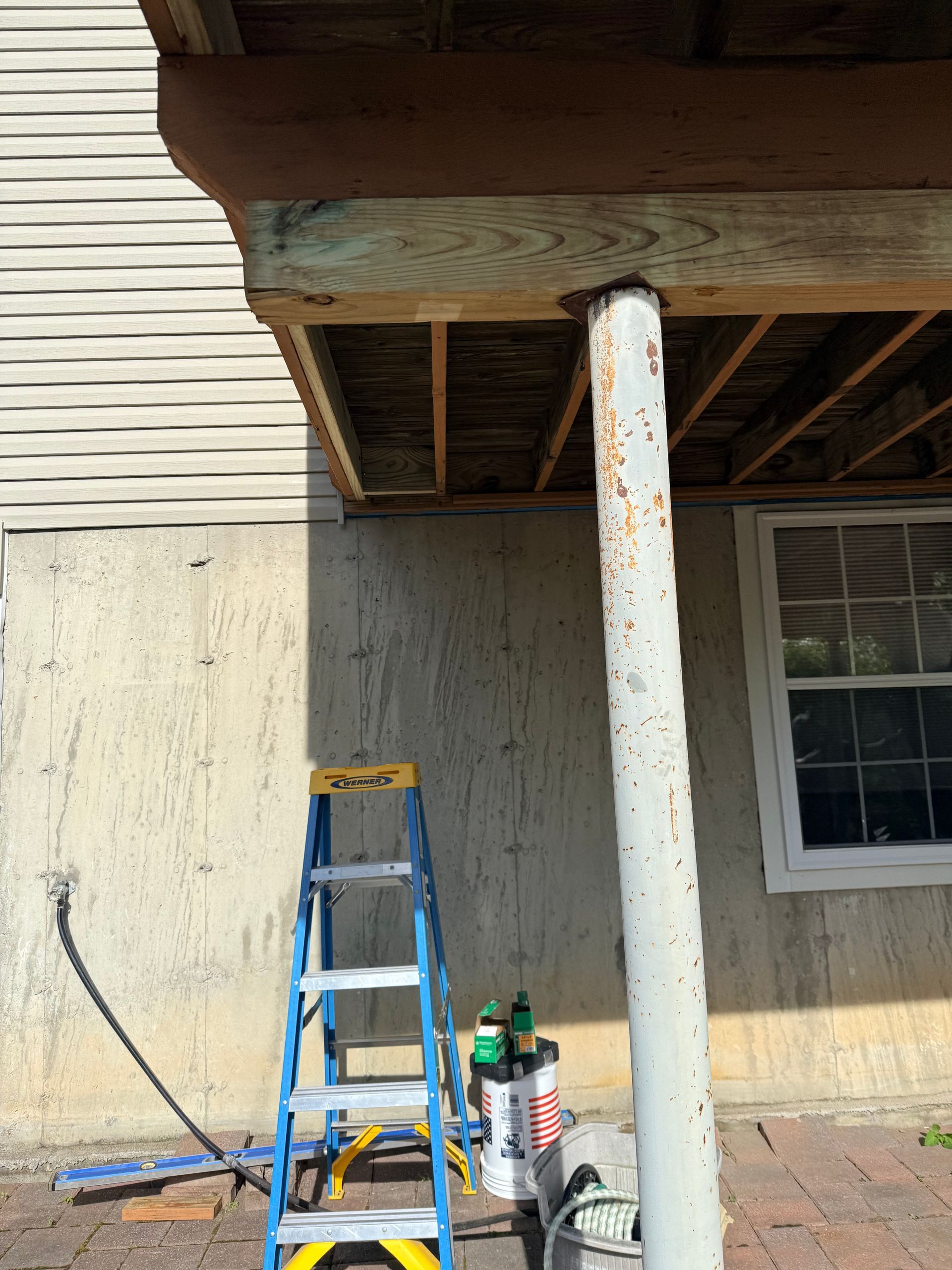 Blue ladder and paint supplies beside a white porch column under a house overhang.