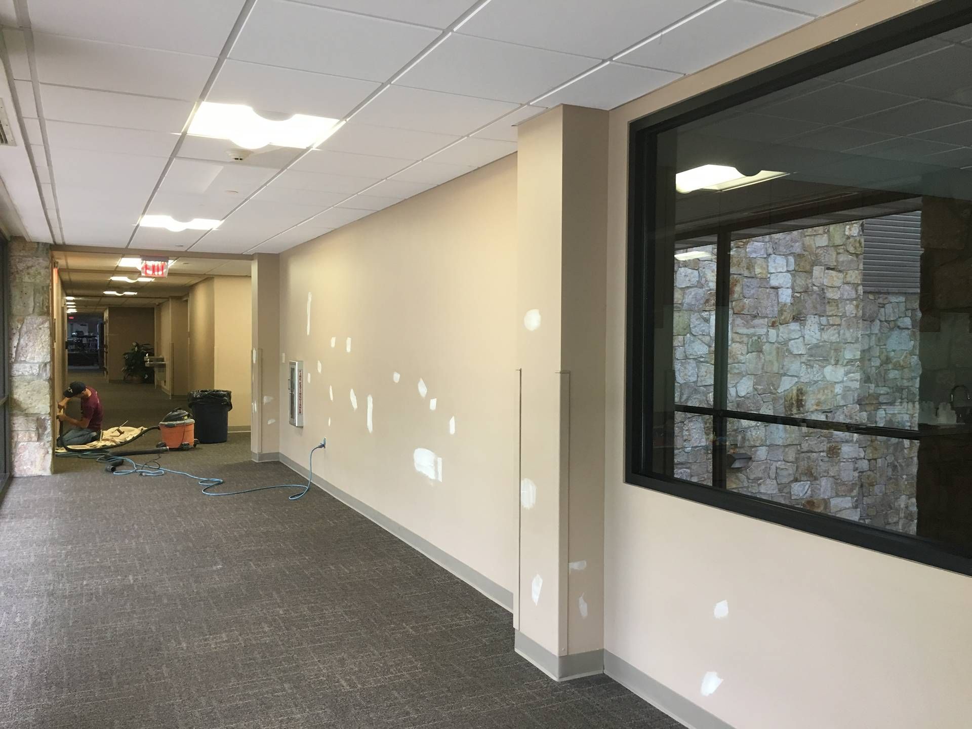 Empty office hallway with beige walls, gray carpet, and a large window on the right.