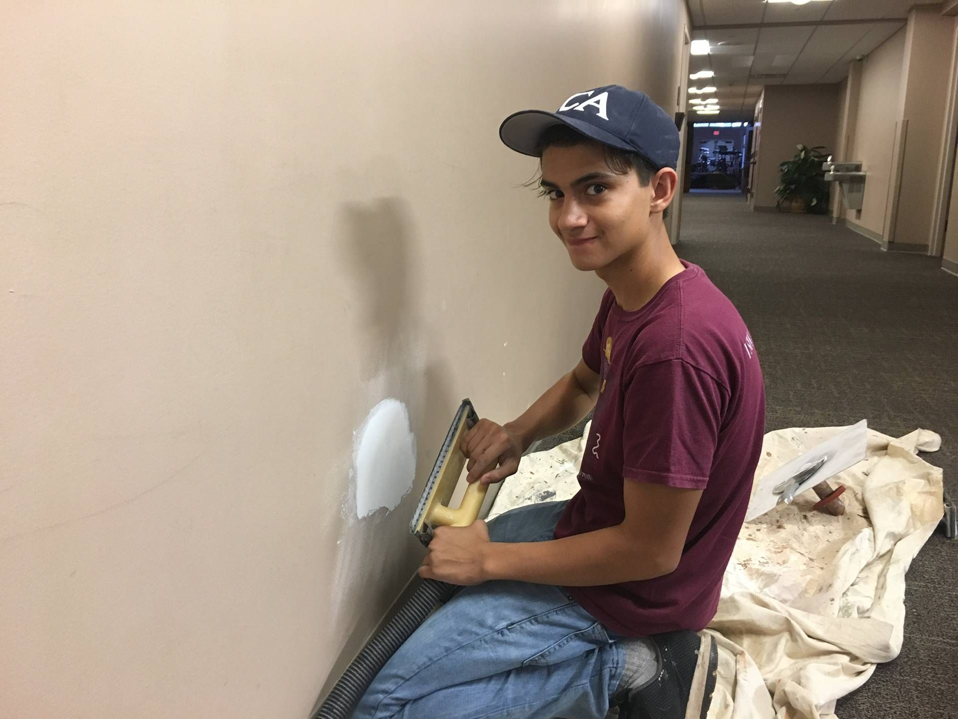 Person kneeling by a wall, painting a white patch with a trowel in a hallway; drop cloth nearby.