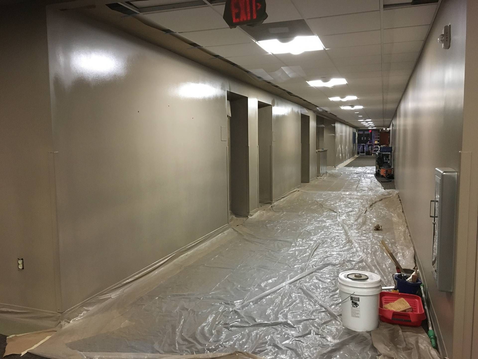 Long hallway under renovation, with plastic-covered floor, beige walls, ceiling lights, and cleaning supplies.