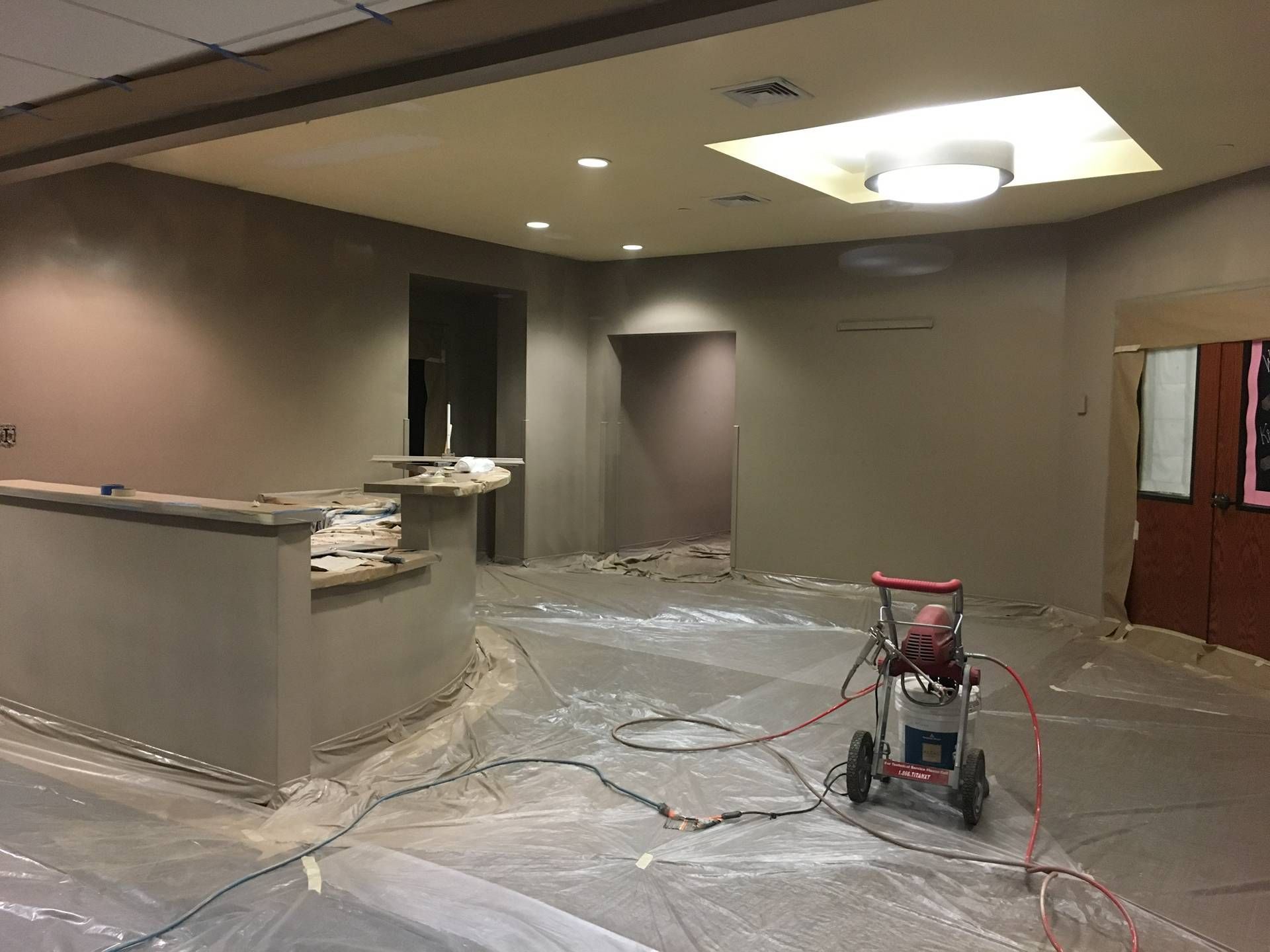Empty room under renovation with covered floor, gray walls, and a paint sprayer on a plastic sheet.