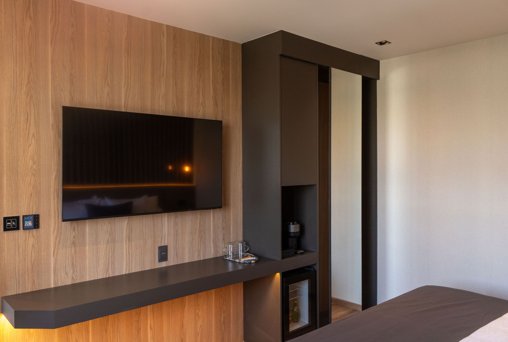 Una moderna habitación de hotel con un televisor de pantalla plana en una pared revestida de madera, una estantería flotante oscura y un minibar integrado.