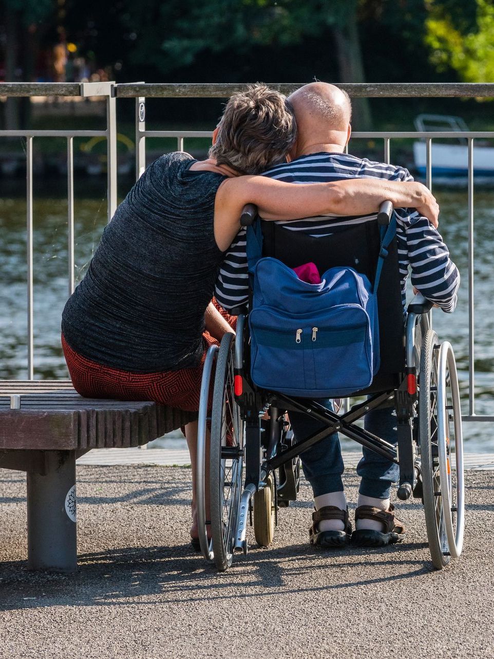 Woman hugs person in wheelchair, both looking at water. They are by a railing and dock.