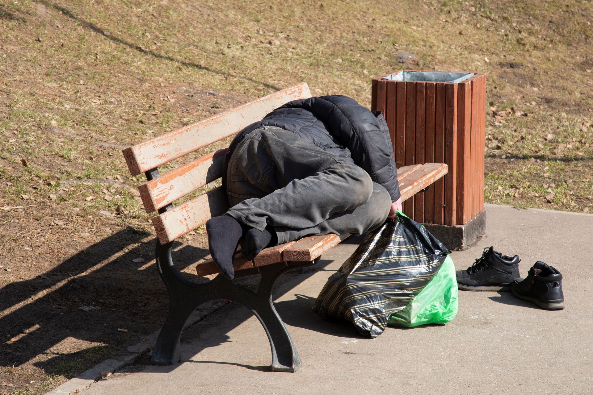 Person sleeping on a park bench, next to trash can and bag; shoes nearby.