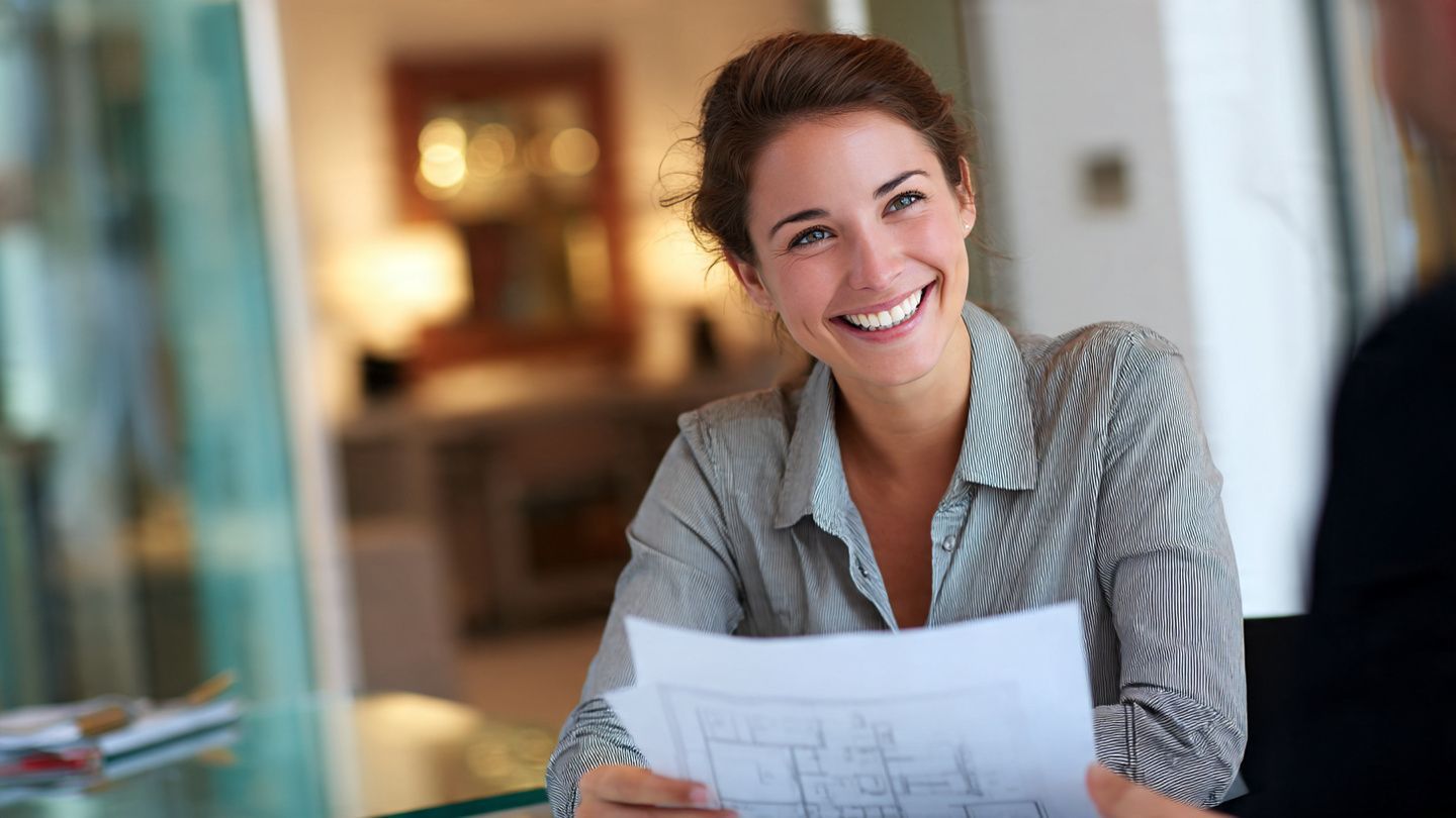 Smiling person in a gray shirt holding papers at a table in a bright office