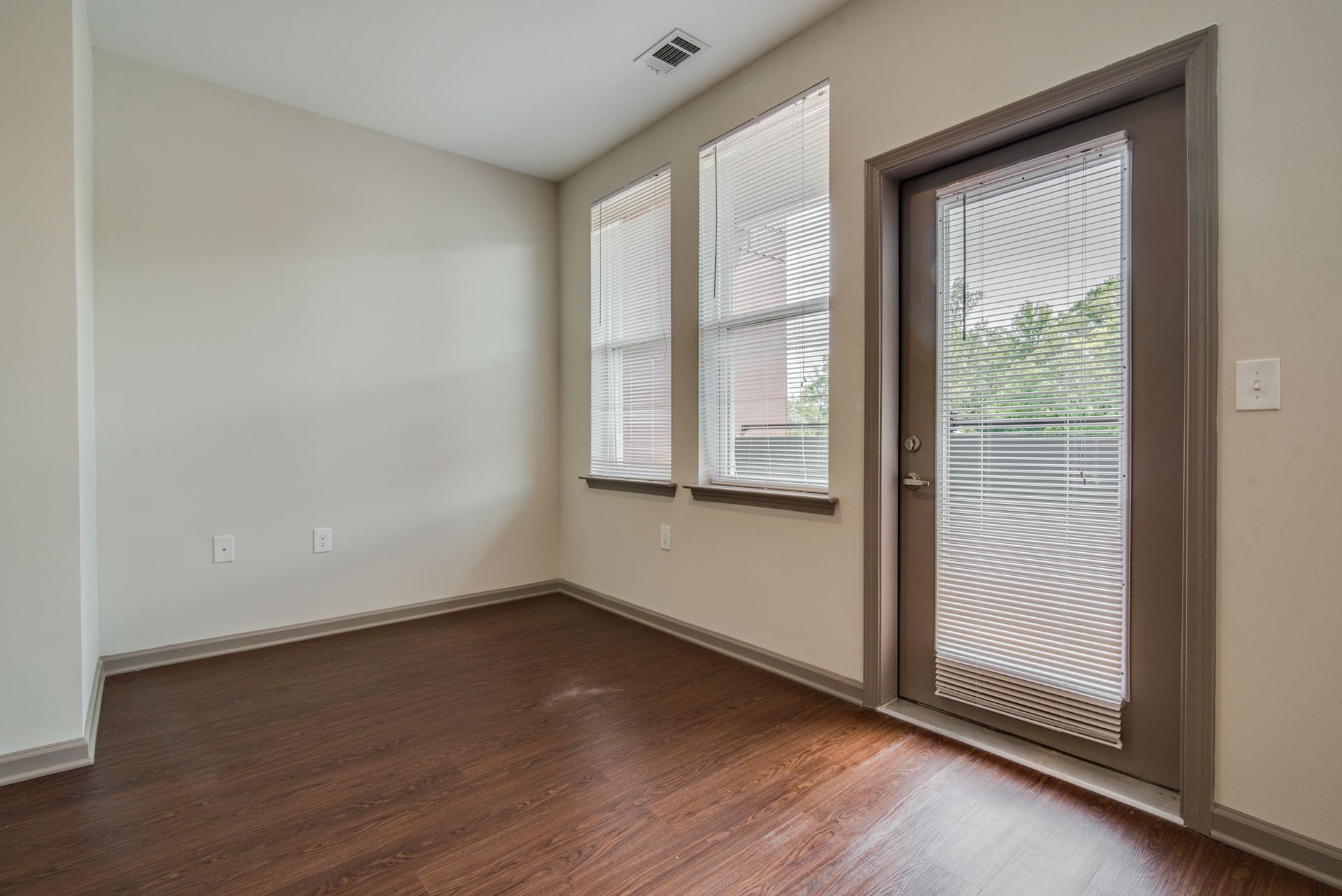 Empty room with dark wood floors, two windows, and a door leading to a balcony at Marvel Apartments.