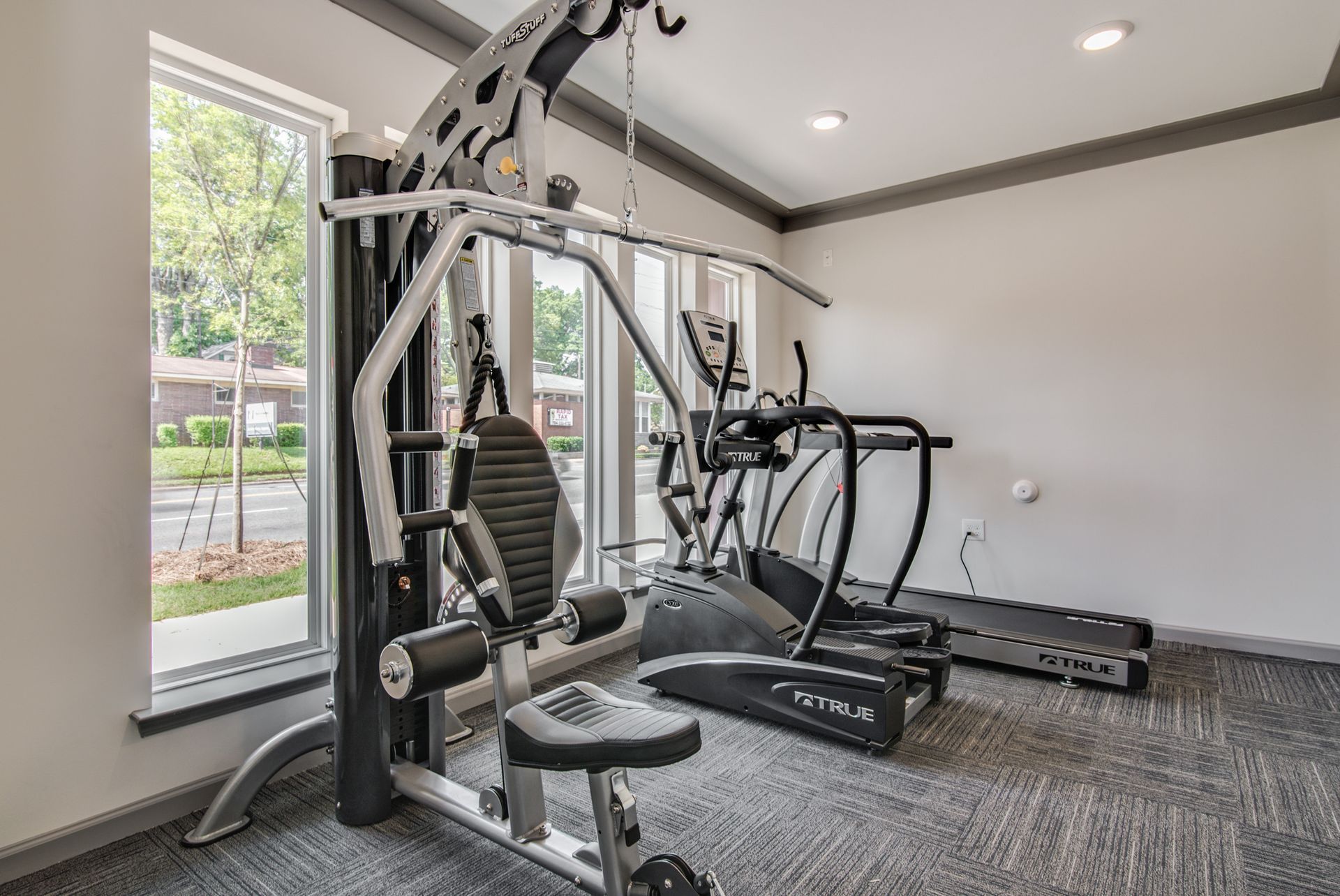 Gym with weight machine, treadmill, elliptical, and window. Gray walls, carpet; fitness equipment at Marvel Apartments.