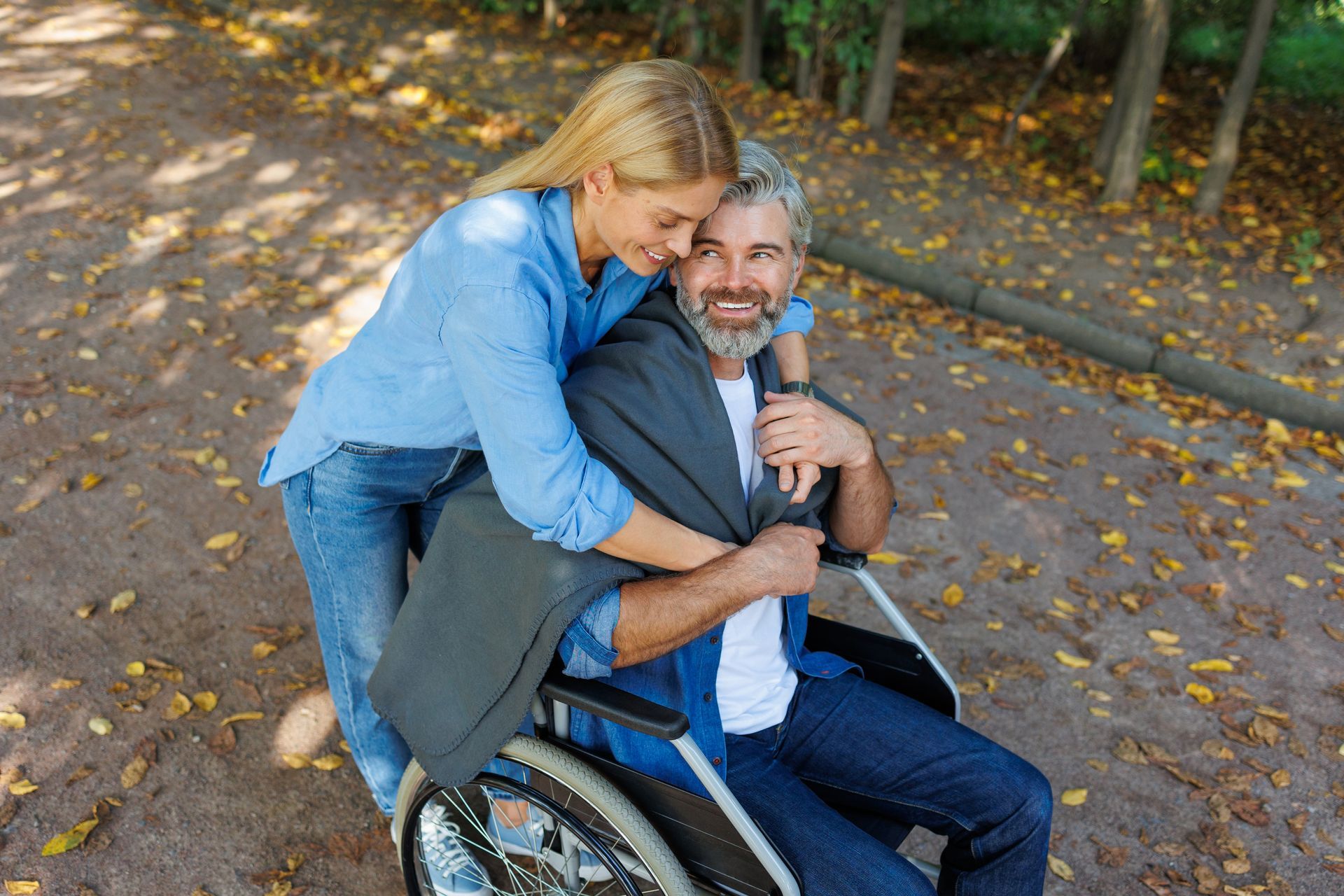 Smiling woman hugging a man in a wheelchair on a leafy park path
